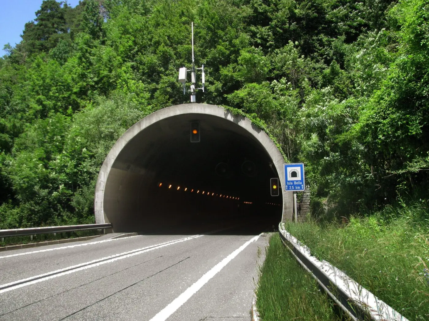 Tunnel che si apre in una zona verdeggiante, con un segnale stradale che indica la distanza. Le luci interne del tunnel sono visibili, mentre l'uscita è in ombra.