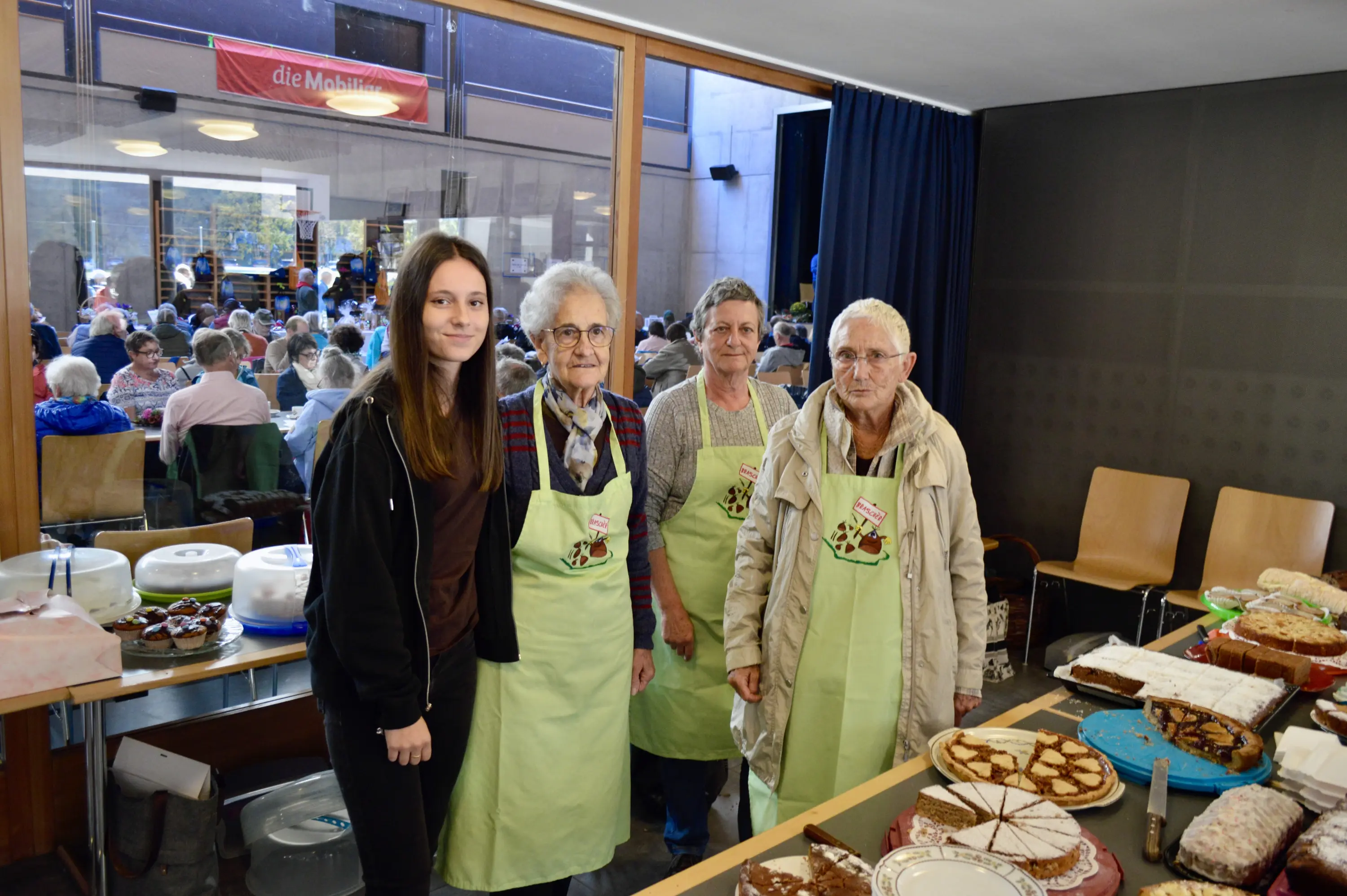 Un gruppo di quattro persone sorridenti, tra cui una giovane donna e tre anziane, in cucina con grembiuli verdi, posano accanto a un tavolo ricoperto di dolci vari. Sullo sfondo, si vede una sala affollata di persone sedute.