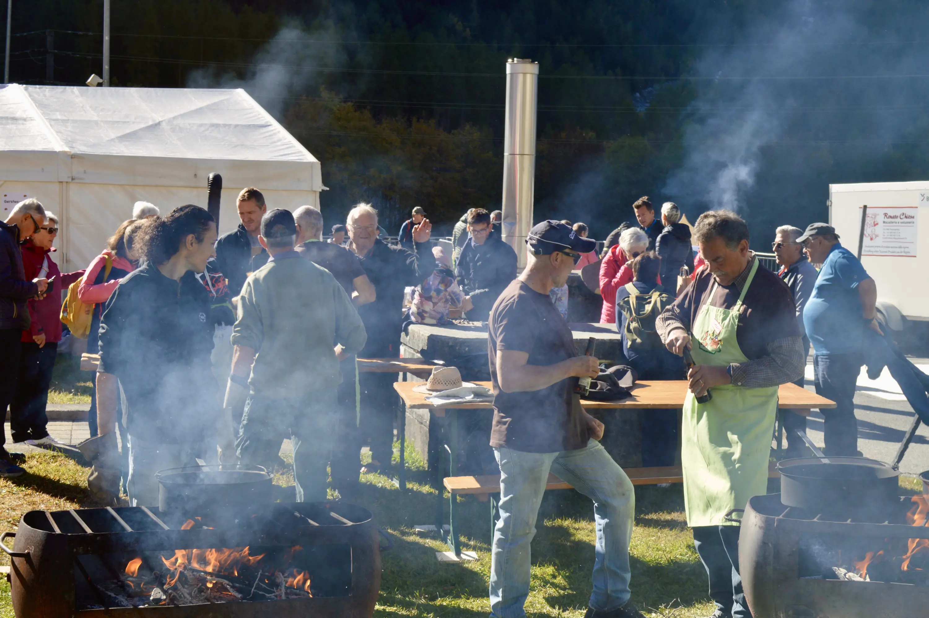 Un gruppo di persone all'aperto partecipa a un evento culinario. Alcuni cuochi preparano cibo su fuochi vivi, mentre altri assaporano le pietanze. Si vede un'atmosfera vivace, con fumi di cottura che si sollevano nell'aria.