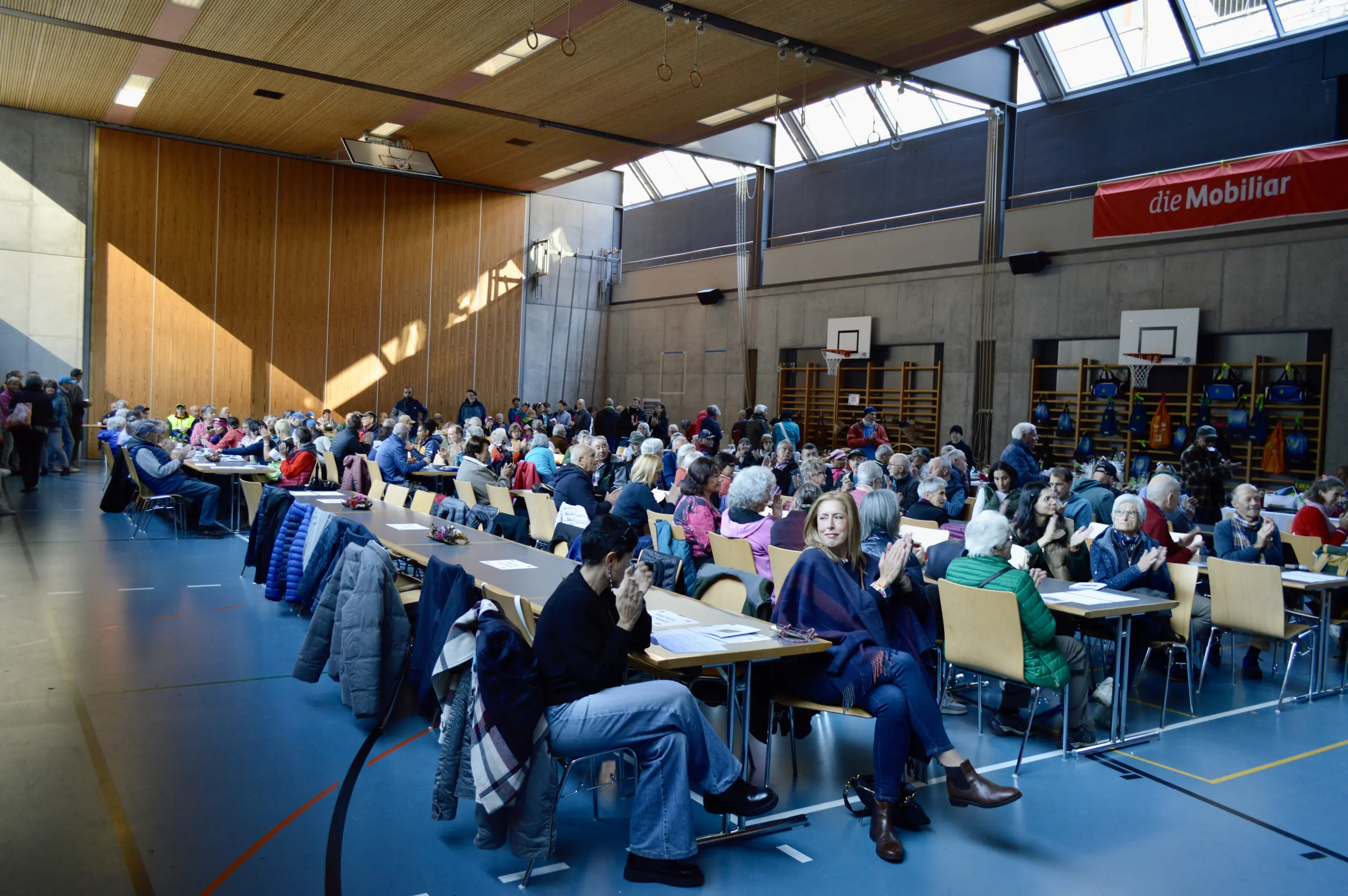 Una grande sala con molte persone sedute ai tavoli, alcune stanno conversando e altre scrivendo. Il luogo è luminoso, con pareti in cemento e grandi finestre. Sullo sfondo ci sono attrezzature sportive e pannelli informativi.