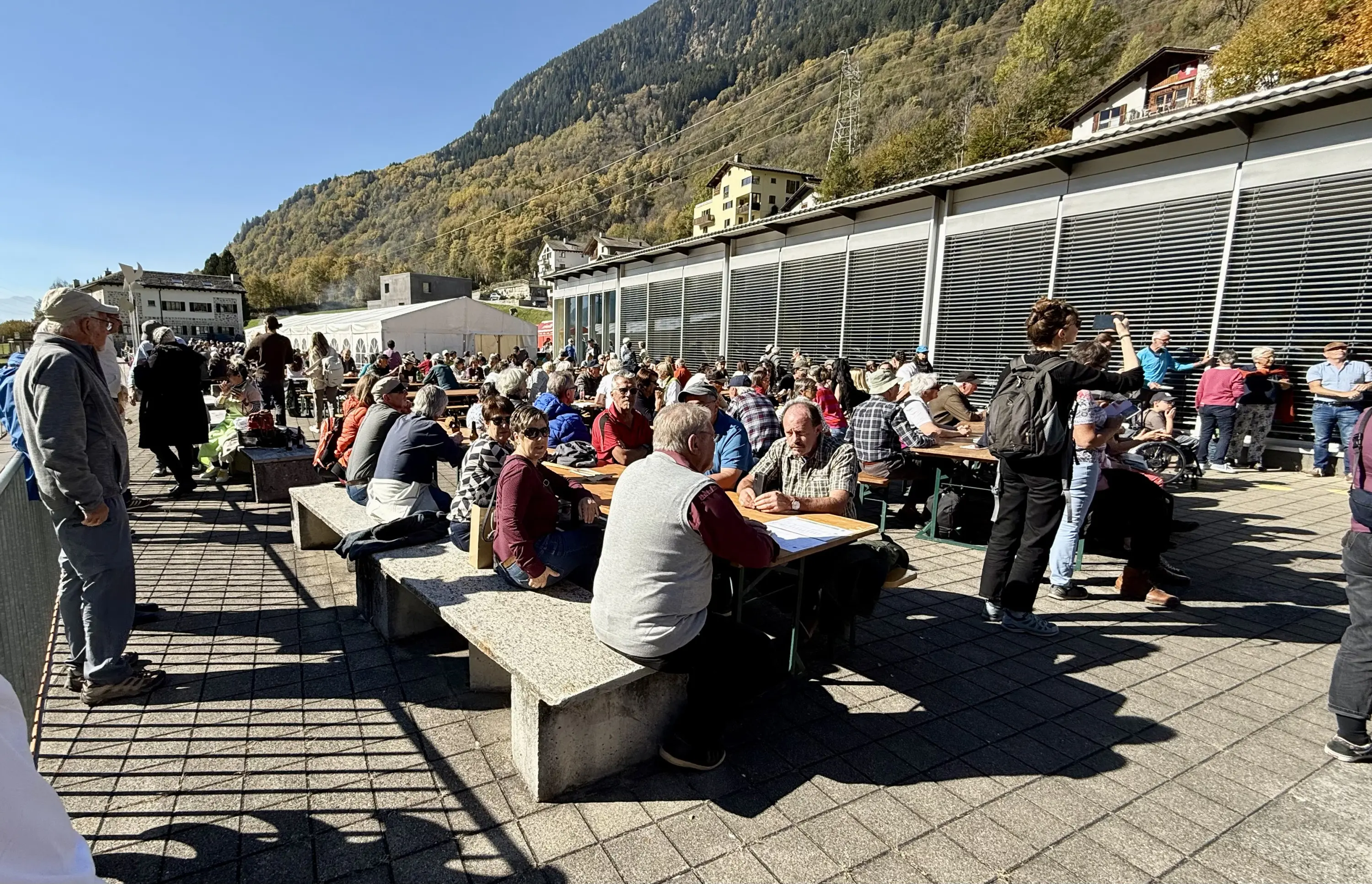 Una grande folla di persone riunite in un'area all'aperto, con tavoli e panchine, circondate da montagne autunnali. Molti partecipanti interagiscono e alcuni alzano le mani.