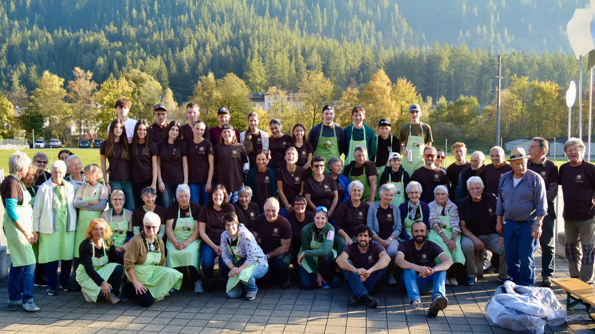 Gruppo di persone sorridenti, vestite con magliette nere e grembiuli verdi, riunite in un campo all'aperto circondato da alberi e montagne.