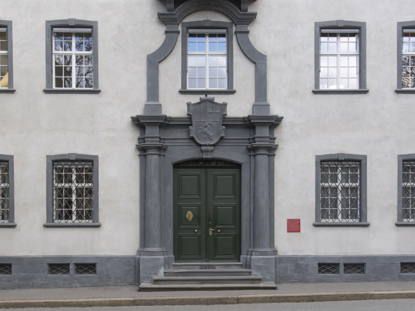 Facciata di un edificio con una porta d'ingresso verde scuro incorniciata da decorazioni grigie. Sopra la porta è presente uno stemma e ai lati ci sono finestre con grate. La parete è di colore chiaro e c'è una targa rossa a destra.