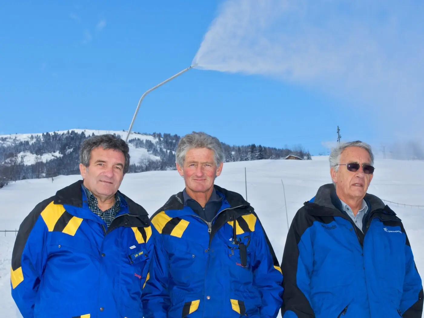 Tre uomini in giacche blu e gialle sorridono in un paesaggio innevato. Sullo sfondo, si vede un impianto che emette vapore nel cielo blu. Gli uomini sono in piedi uno accanto all'altro, circondati da un panorama montano.