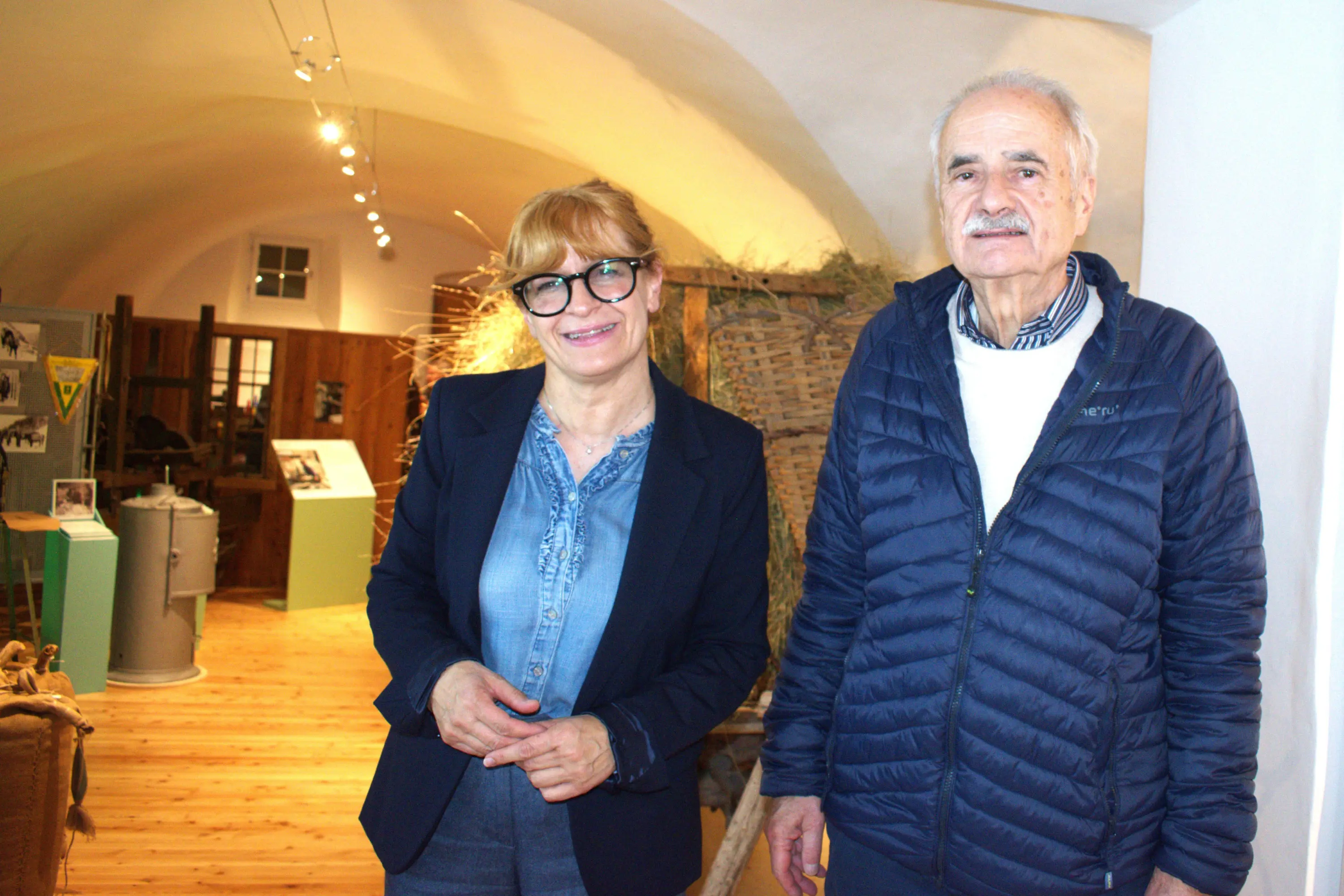 Due persone sorridenti in un museo. La donna indossa occhiali grandi e una giacca nera, mentre l'uomo ha i capelli grigi ed è vestito con un giubbotto blu. Sullo sfondo si vedono oggetti esposti.