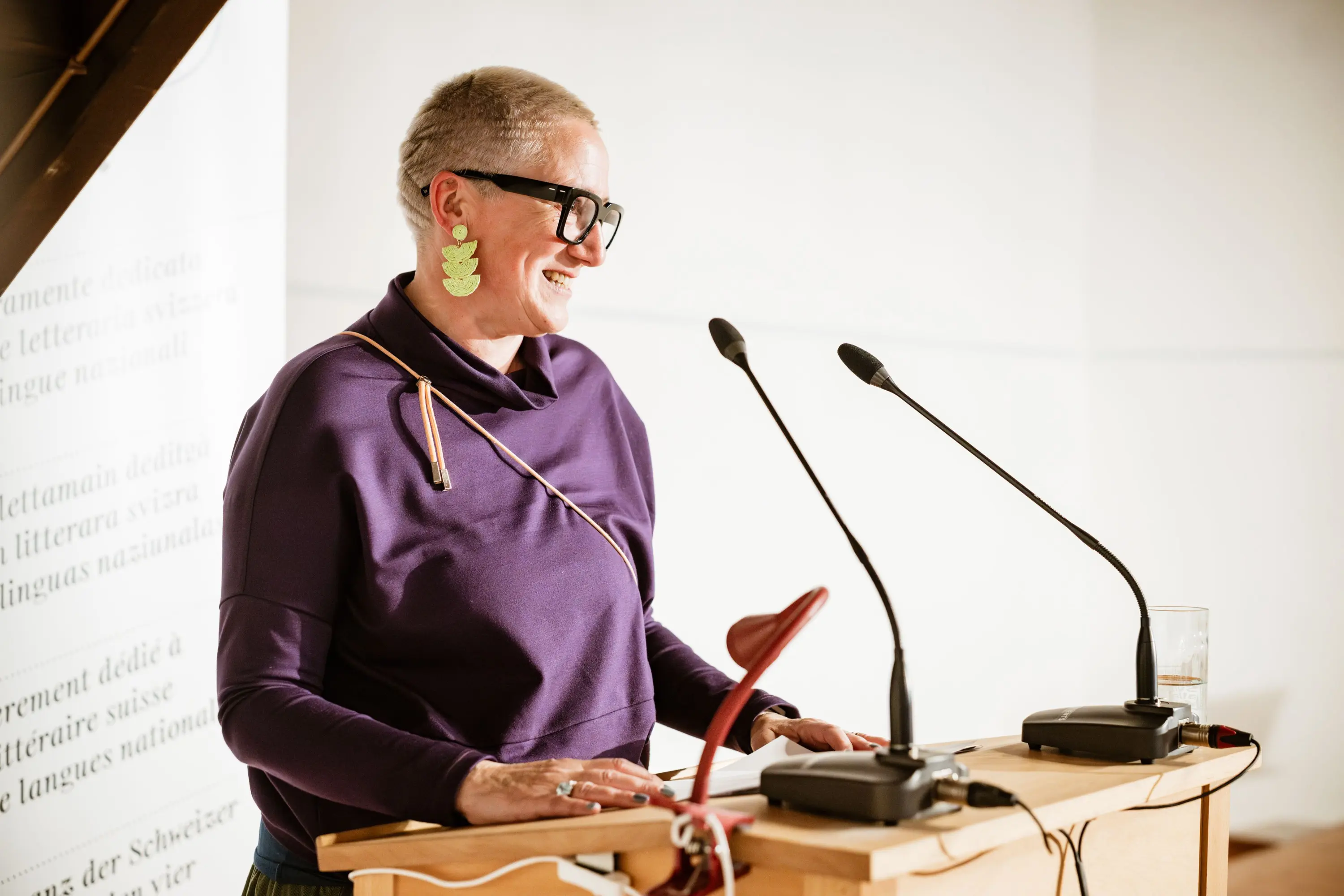 Una persona al microfono, sorridente, indossa occhiali neri e orecchini verdi, mentre si prepara a parlare da un leggio con due microfoni.