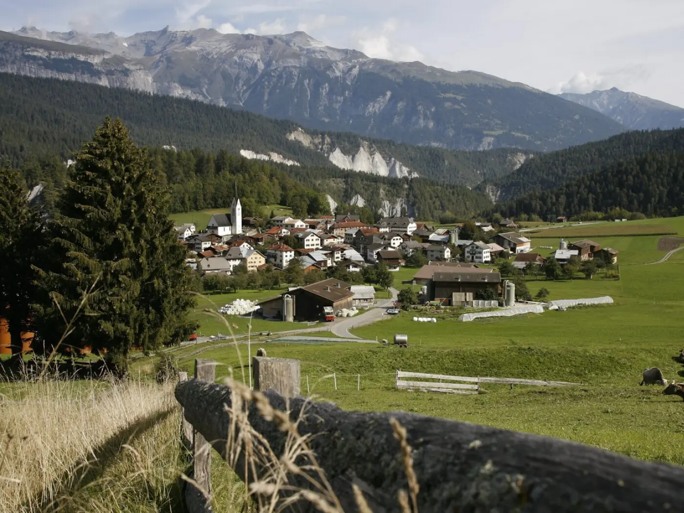 Un paesaggio rurale con un villaggio circondato da montagne. Abitazioni sparse, una chiesa bianca e campi verdi. Un lungo recinto di legno in primo piano.