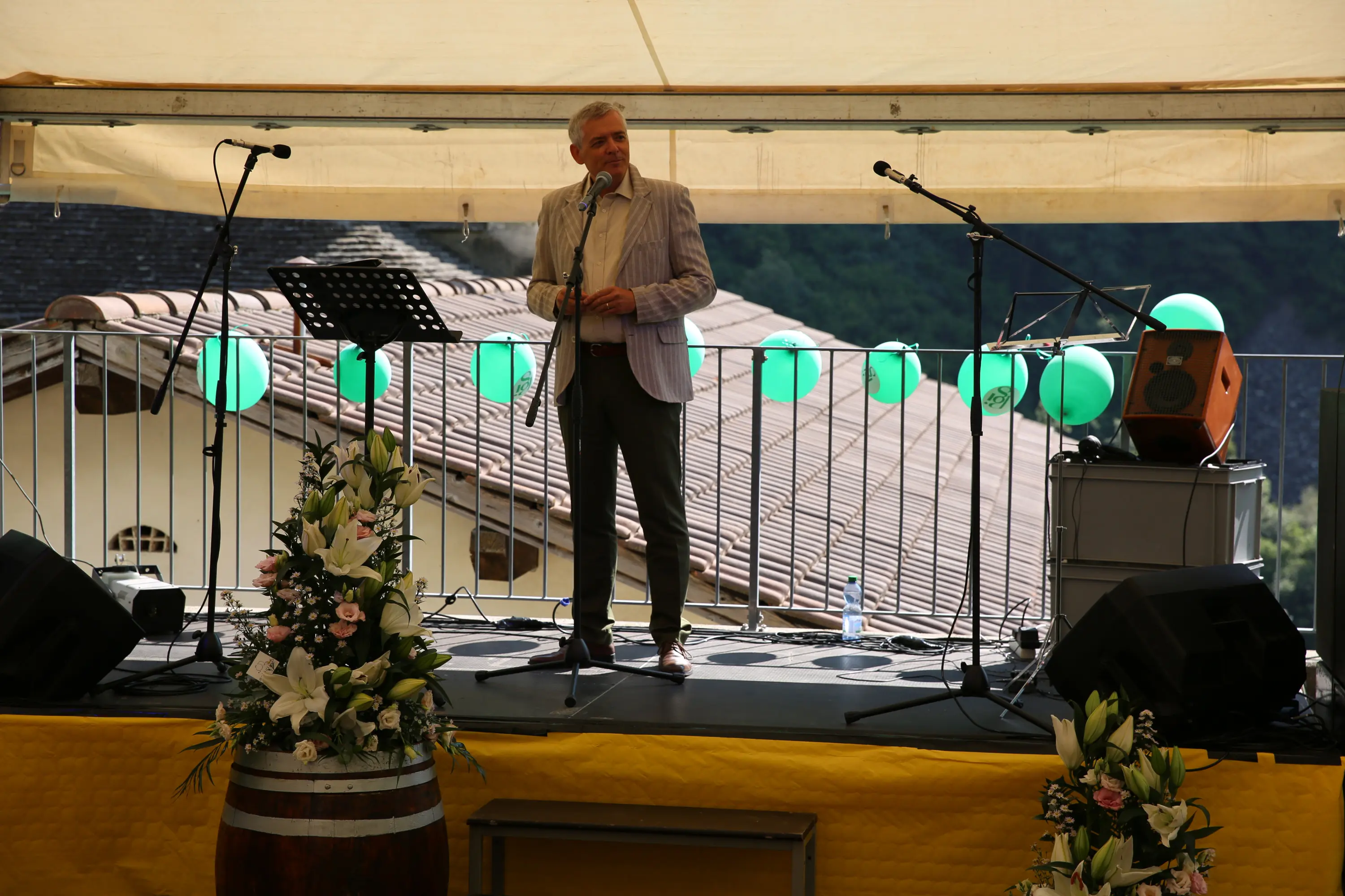 Un uomo in giacca e cravatta parla a un microfono su un palco, con strumenti musicali e decorazioni verdi sullo sfondo. Barili e fiori adornano il palco.