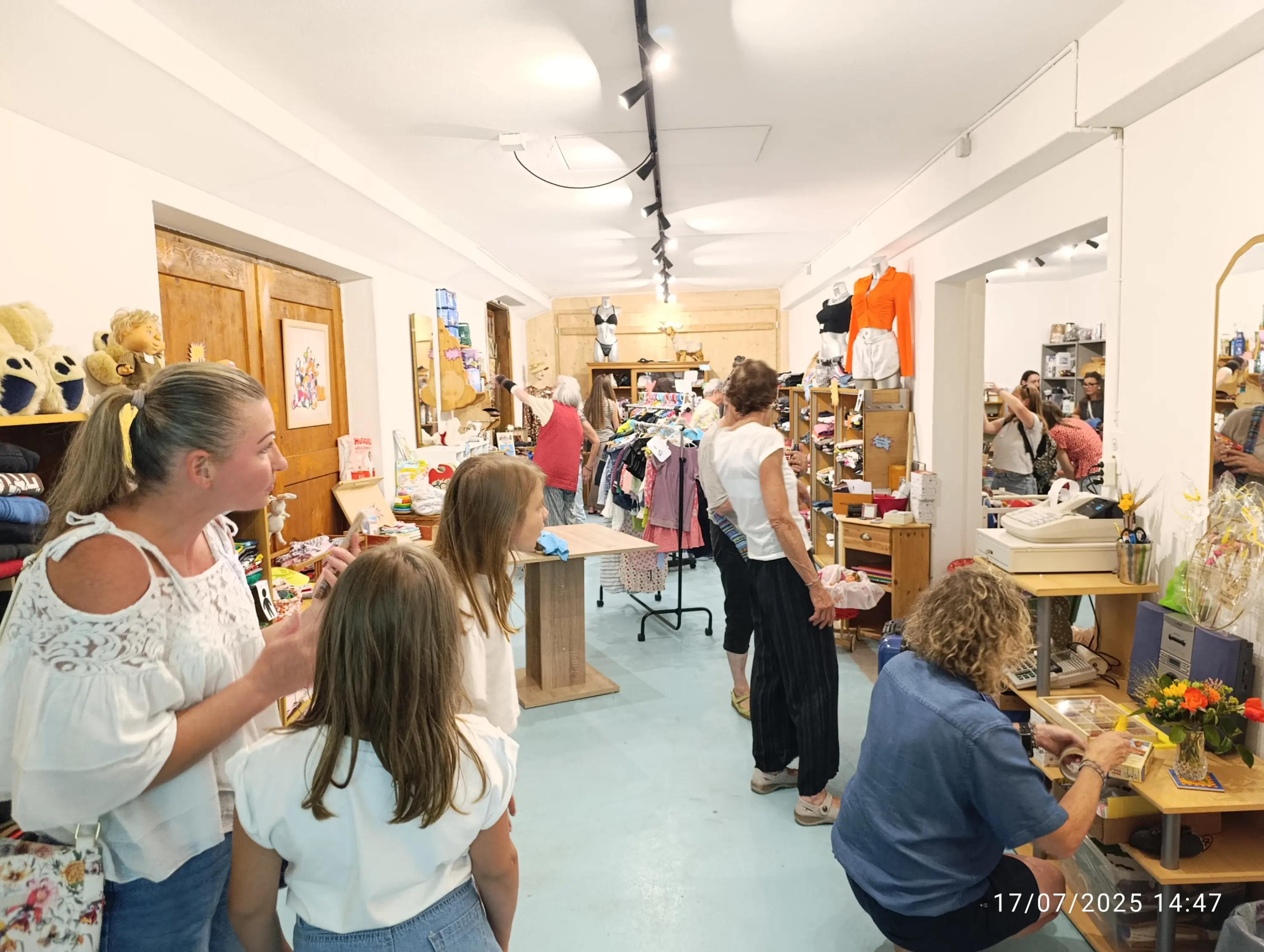 Un negozio di artigianato con persone che interagiscono. Due donne chiacchierano mentre una ragazza osserva. In fondo, una donna lavora a un tavolo e altre persone amministrano il negozio. Scaffali pieni di oggetti vari.