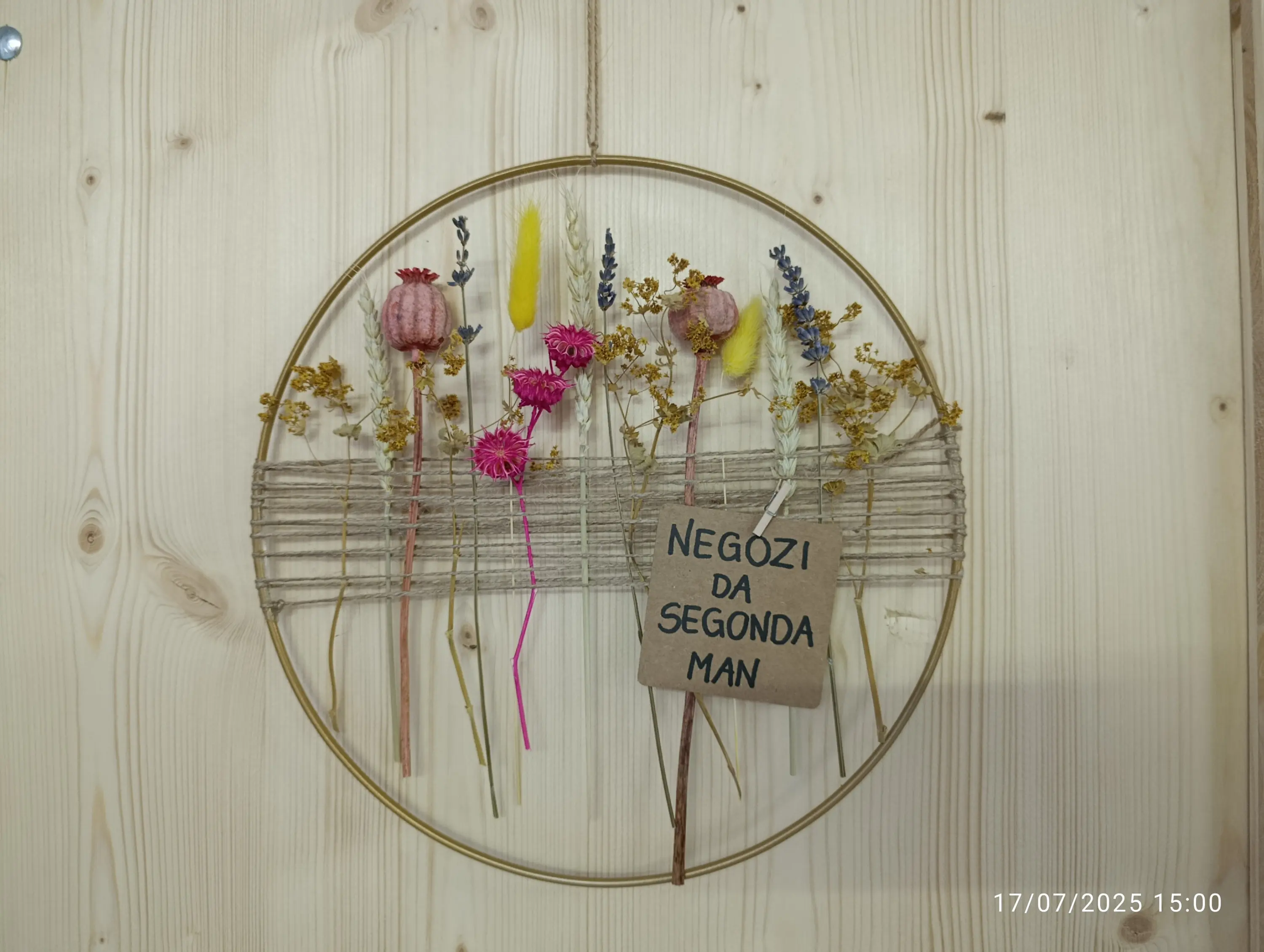 Una decorazione rotonda con fiori secchi colorati, inclusi papaveri e lavanda, fissata su una struttura di legno. Un cartellino con la scritta "NEGOZI DA SECONDA MAN" è appeso al centro.