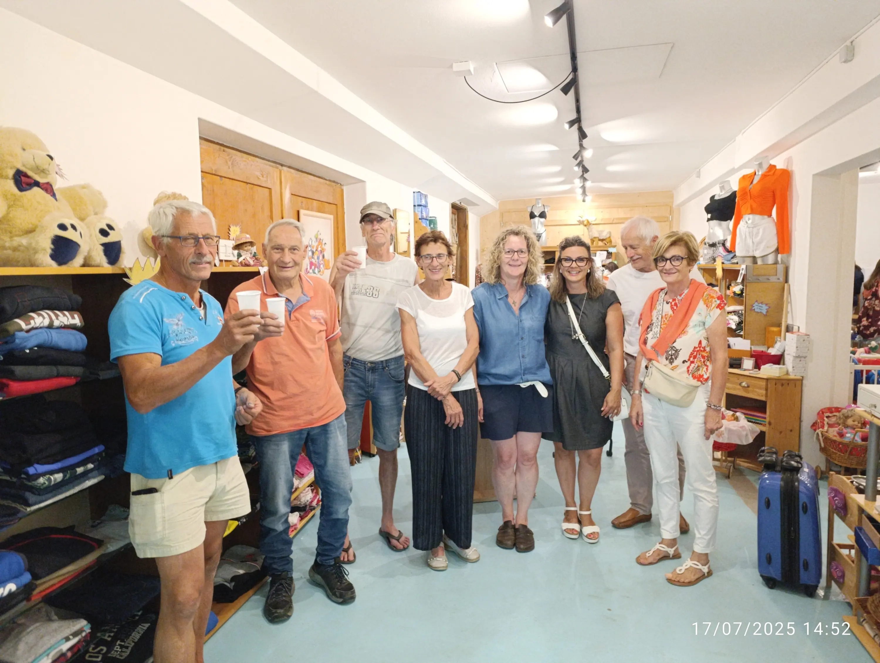 Un gruppo di otto persone sorridenti, in un negozio ben illuminato, mentre fanno un brindisi. Sono vestiti in modo informale e si trovano davanti a scaffali con abbigliamento e peluche.