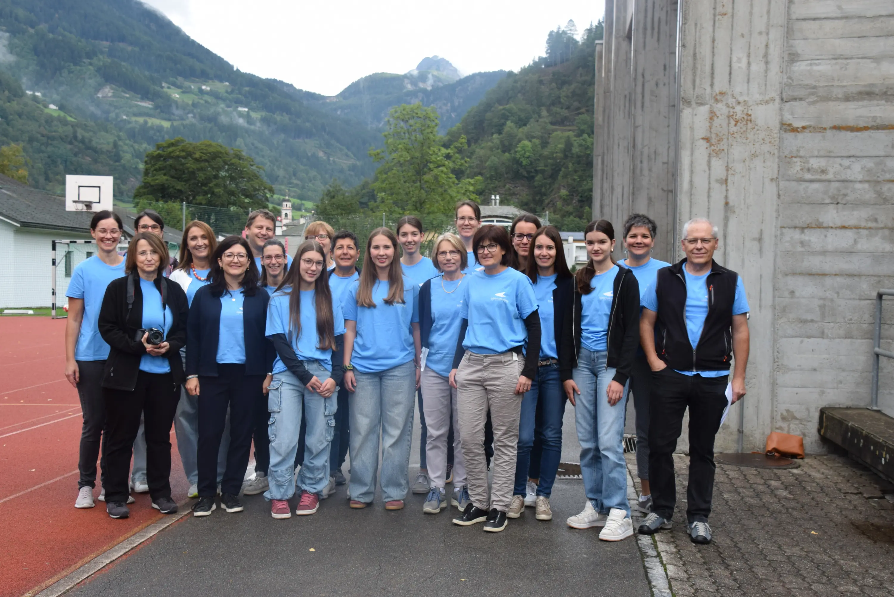 Gruppo di persone vestite con magliette azzurre e pantaloni chiari, sorridenti in un'area all'aperto con montagne sullo sfondo.