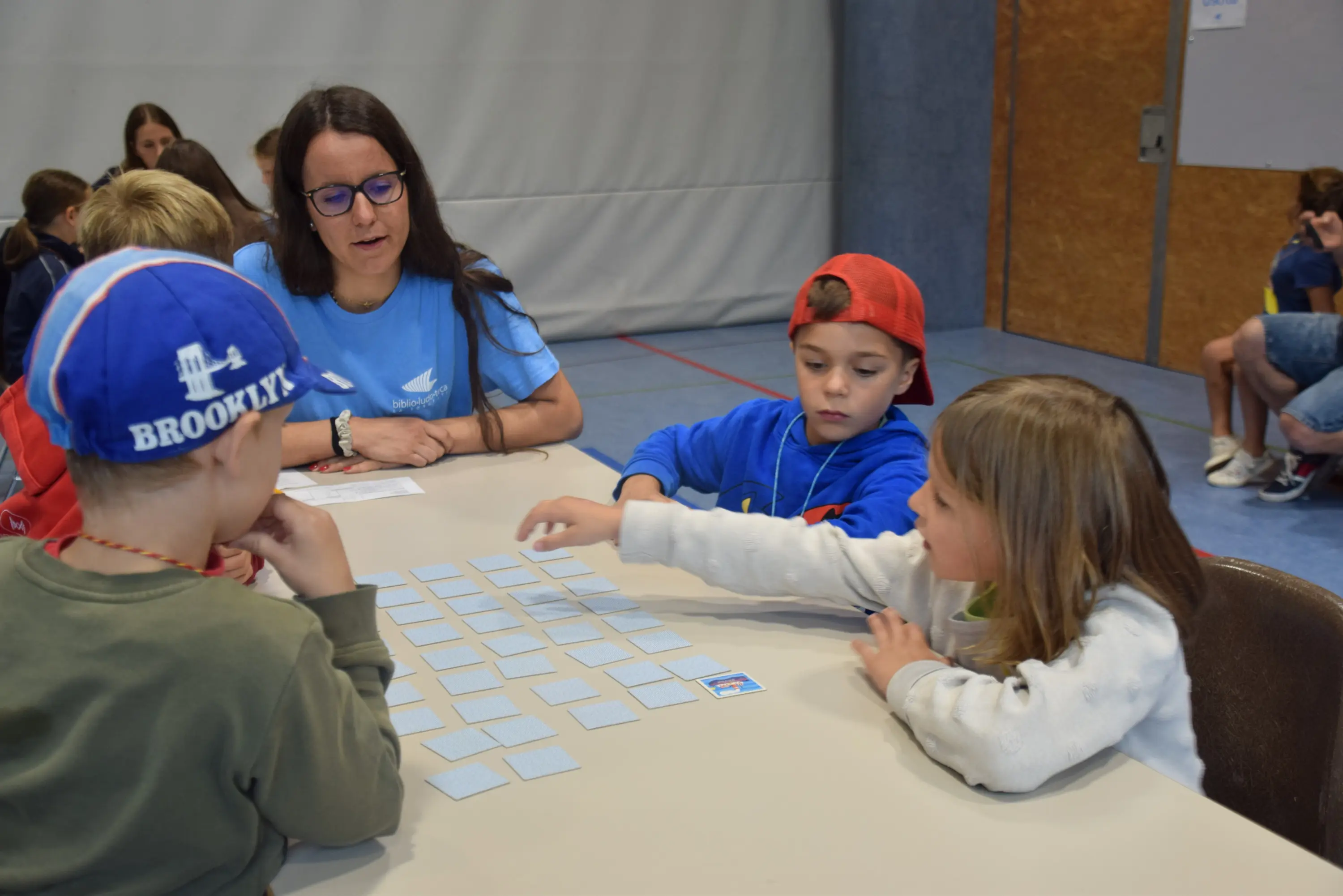 Gruppo di bambini che giocano a un gioco di memoria su un tavolo. Una donna osserva mentre un bambino in maglietta blu e uno in cappellino rosso scelgono delle carte. Un'altra bambina, con i capelli biondi, indica una carta.