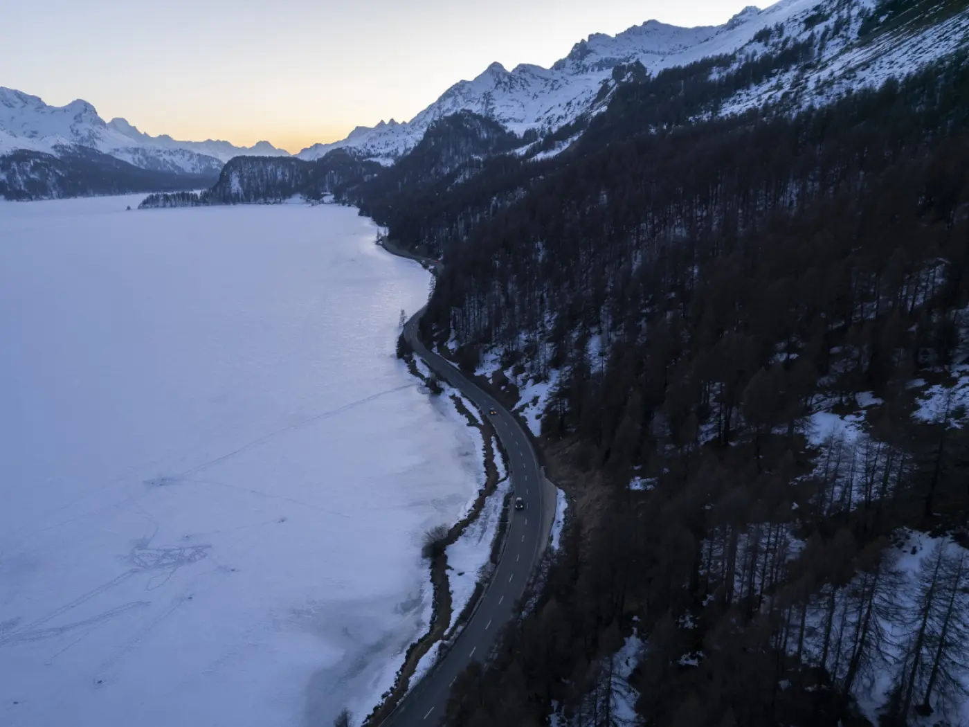 Panorama di un lago ghiacciato circondato da montagne innevate. Una strada si snoda lungo la riva del lago, con alberi senza foglie sul lato destro. Il cielo presenta tonalità calde al tramonto.