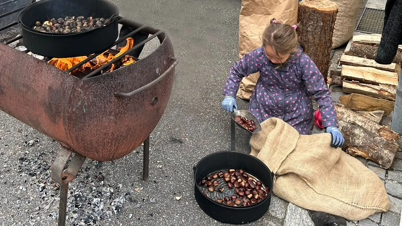 Una bimba con un grembiule e guanti blu raccoglie castagne tostate da un recipiente, mentre un grande bracere in metallo produce fiamme. Sullo sfondo, un altro recipiente contiene castagne.