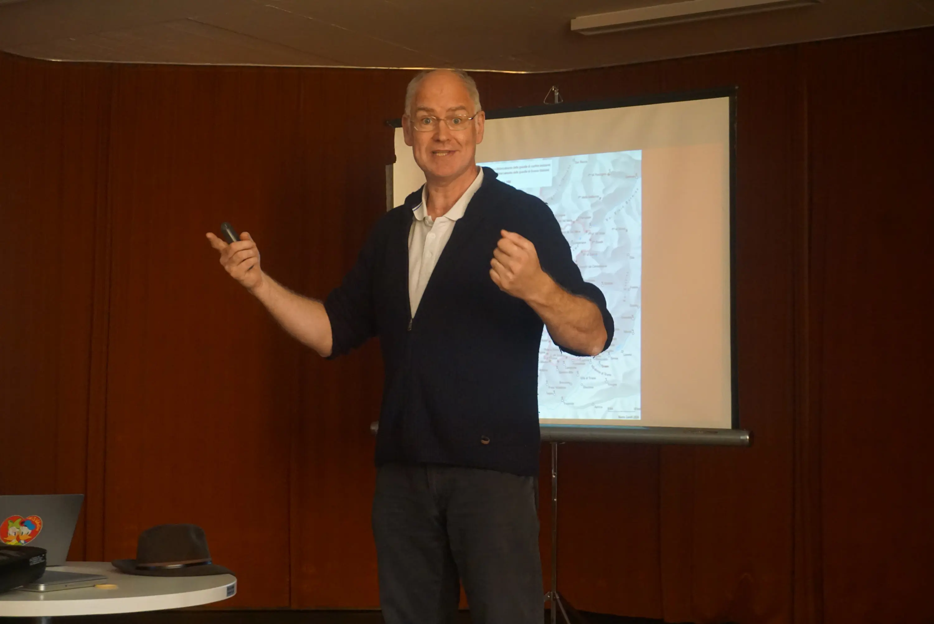 Un uomo con occhiali e una maglia blu scuro sta parlando a un pubblico. Dietro di lui c'è un proiettore che mostra una mappa. La stanza ha pareti color legno e un tavolo con un computer portatile.