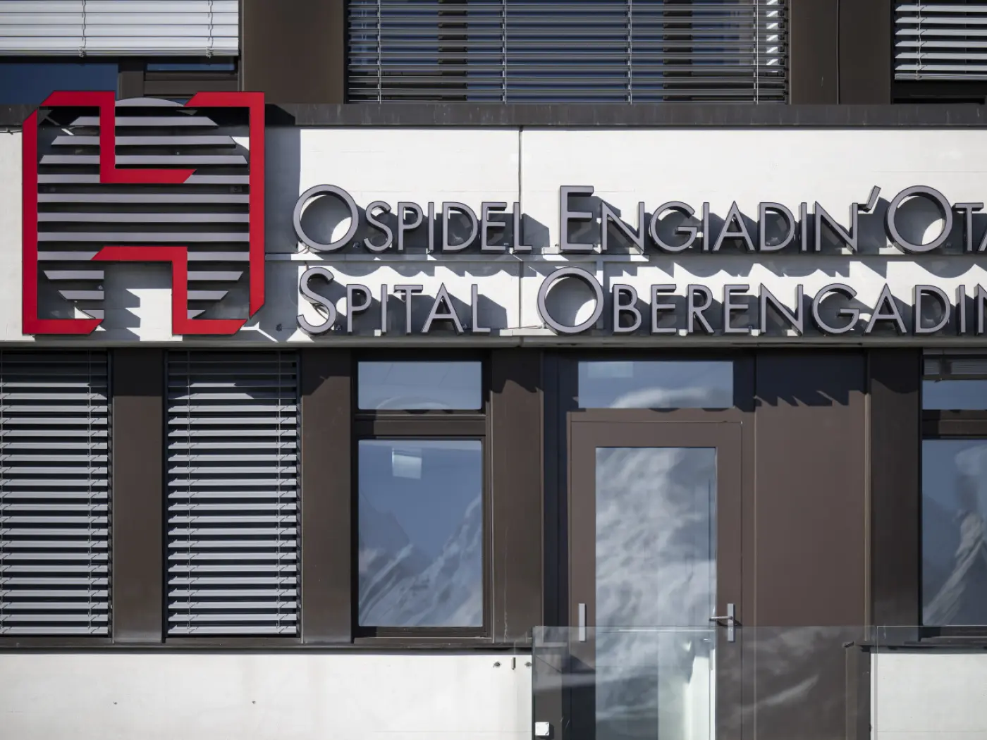 Insegna del "Ospidel Engiadin'Ot" con il logo rosso e testo in italiano e tedesco. La facciata è moderna con finestre a persiane.