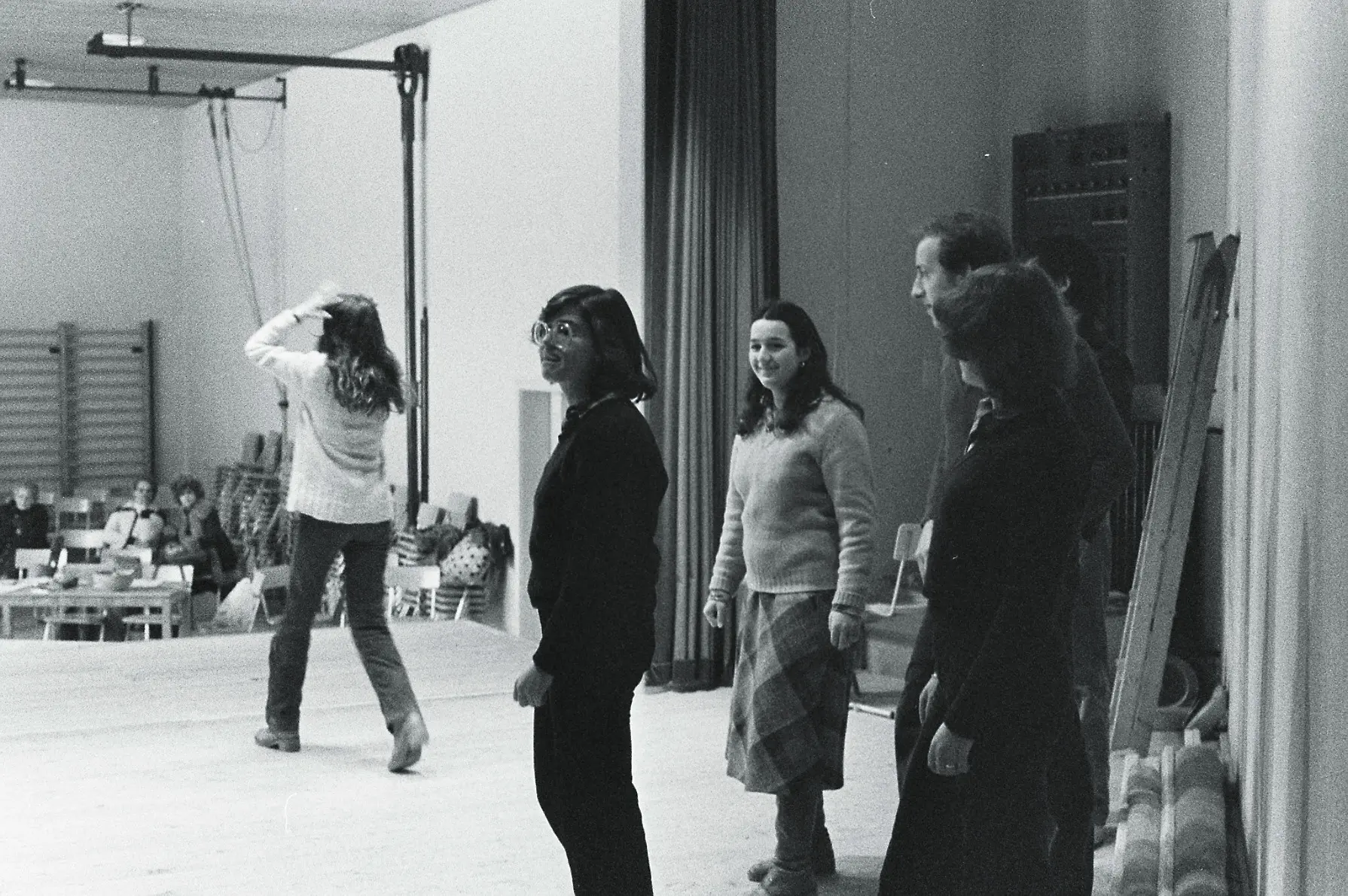 Un gruppo di cinque persone si trova su un palcoscenico, con una donna che si allontana verso il fondo. Due donne sorridono e guardano in avanti, mentre un uomo e un'altra donna sono in secondo piano. L'ambiente è luminoso e ci sono sedie disposte sul fondo.