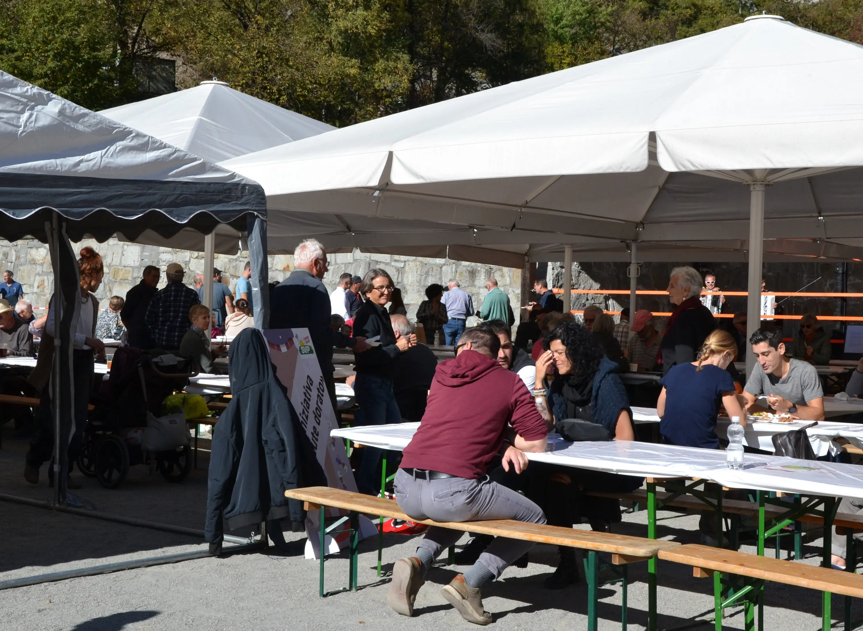 Un gruppo di persone sedute e in piedi sotto tende bianche, mentre mangiano e conversano in un'atmosfera conviviale. Tavoli di legno con cibo e bevande sono visibili, e sullo sfondo si nota una parete in pietra e alberi.