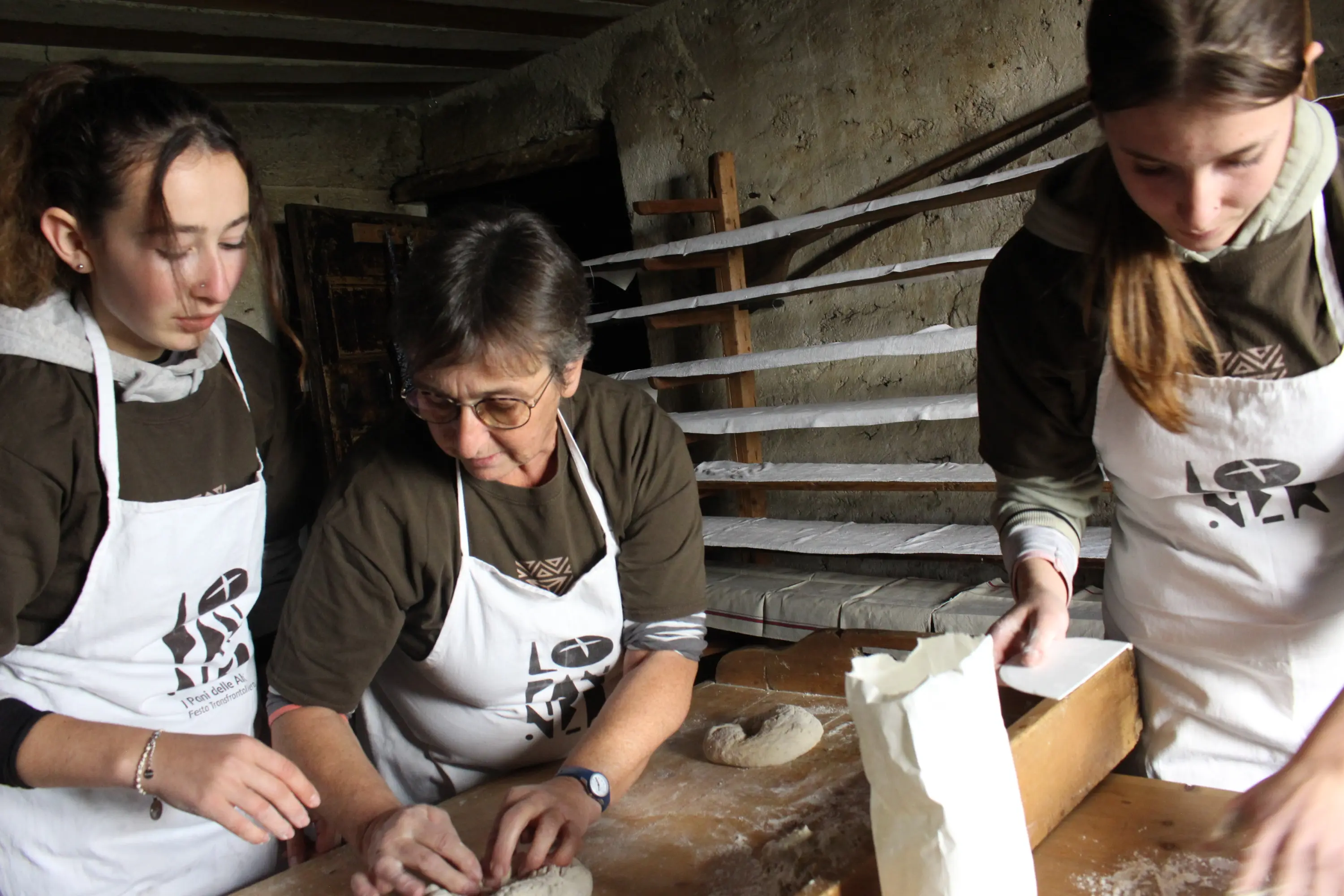 Tre donne si dedicano a fare il pane in una cucina rustica. Indossano magliette marroni e grembiuli bianchi mentre lavorano con la pasta su un tavolo di legno. Sullo sfondo si vedono scaffali con attrezzi da cucina.