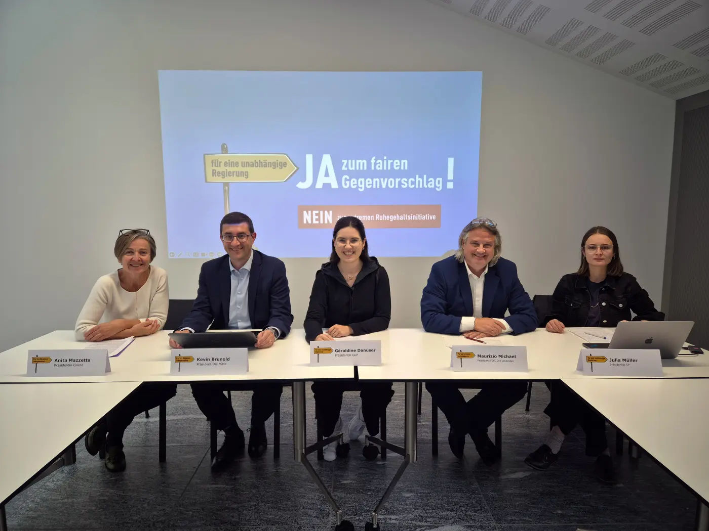 Cinque persone sedute attorno a un tavolo, con un proiettore sullo sfondo che mostra un manifesto. I partecipanti discutono un'iniziativa di governo, con cartelli che dicono "JA zum fairen Gegenvorschlag!" e "NEIN".