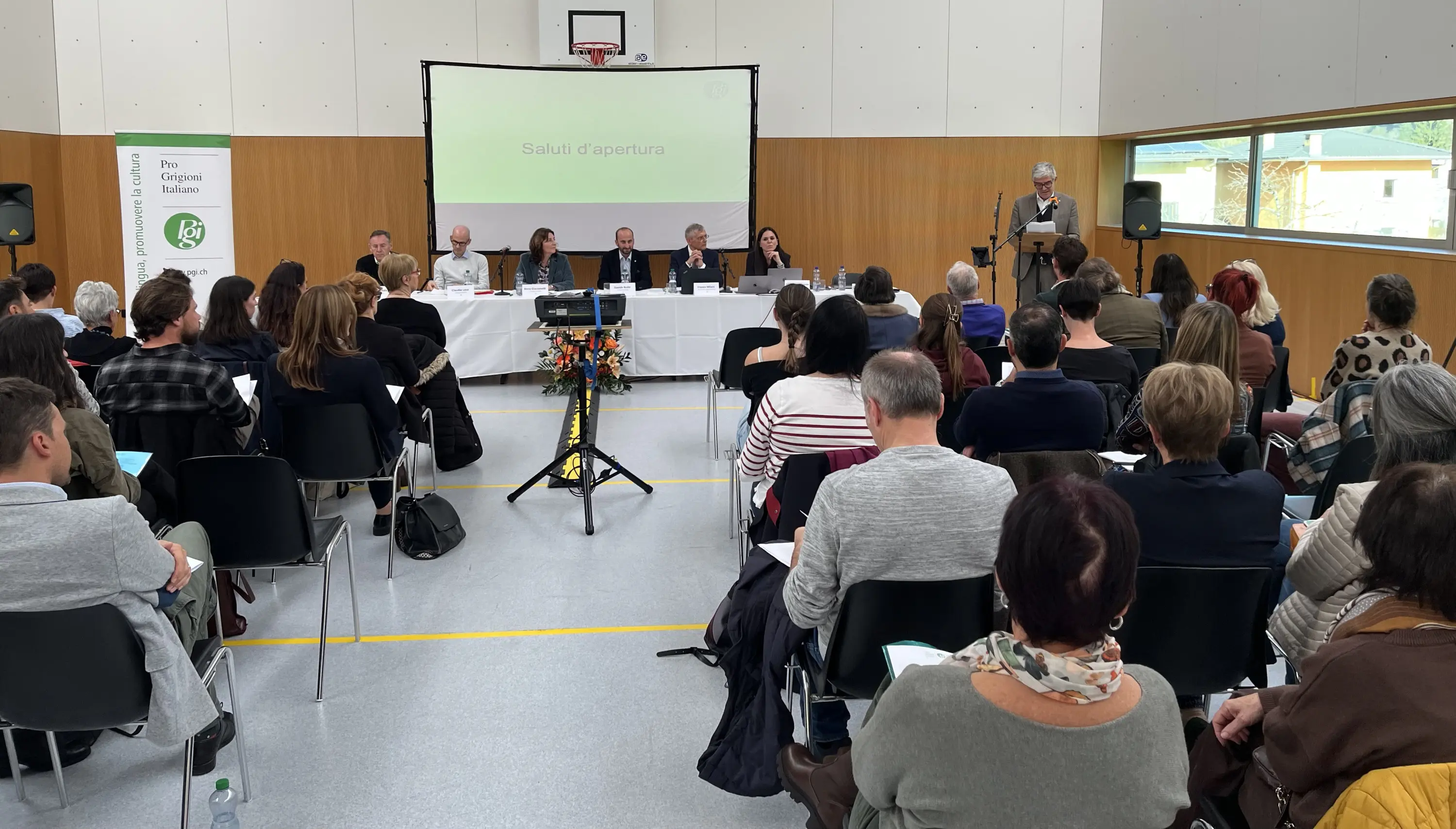 Sala affollata durante un evento, con tavolo dei relatori e una persona che parla in fondo. Sullo sfondo, una presentazione su schermo. Pubblico attento, seduto su sedie nere. Decorazioni floreali sul tavolo.