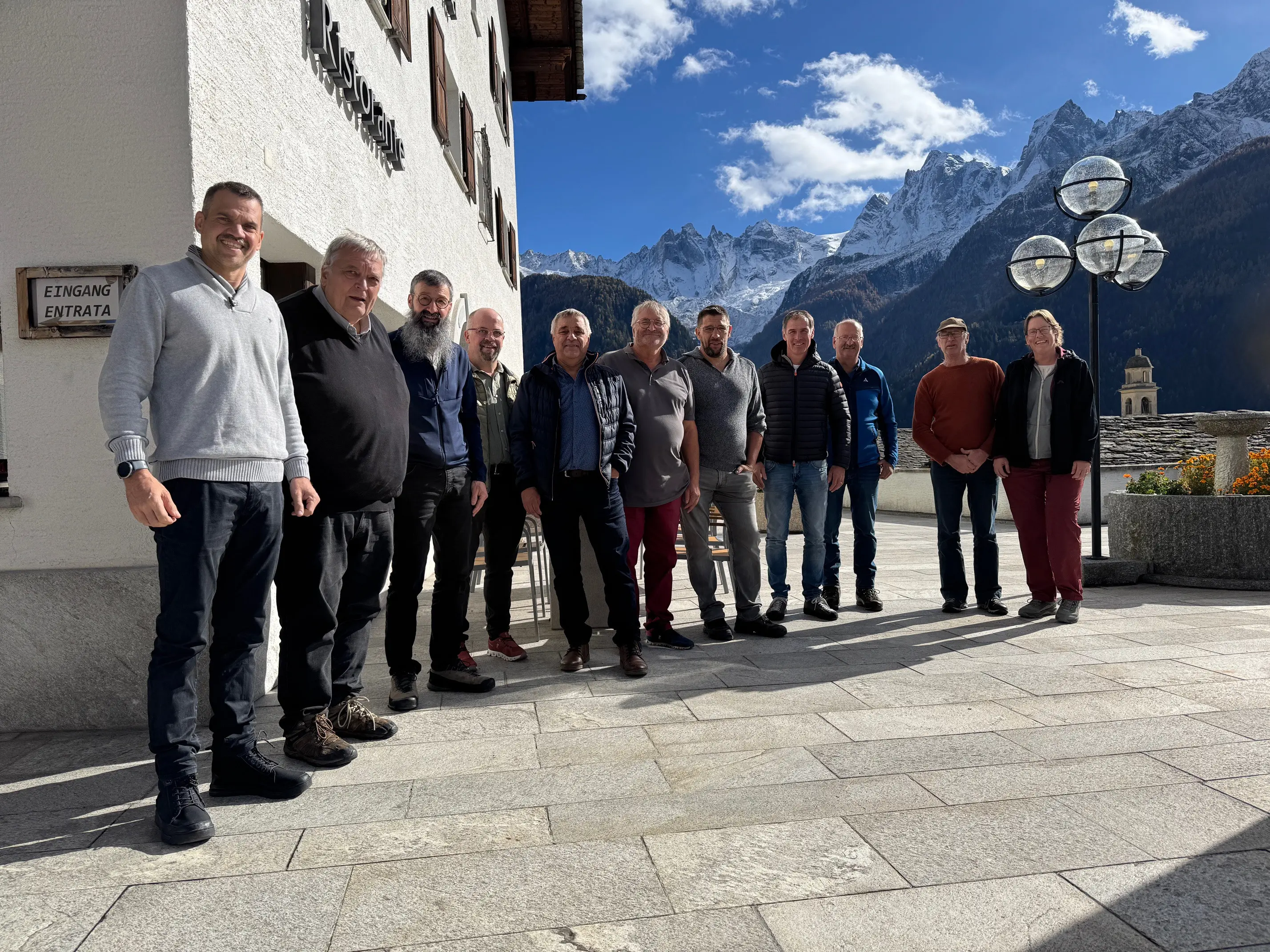 Gruppo di uomini in piedi all'aperto, davanti a una montagna con cime innevate, sorridono verso la fotocamera. Il cielo è blu con nuvole bianche e ci sono edifici in stile alpino sullo sfondo.