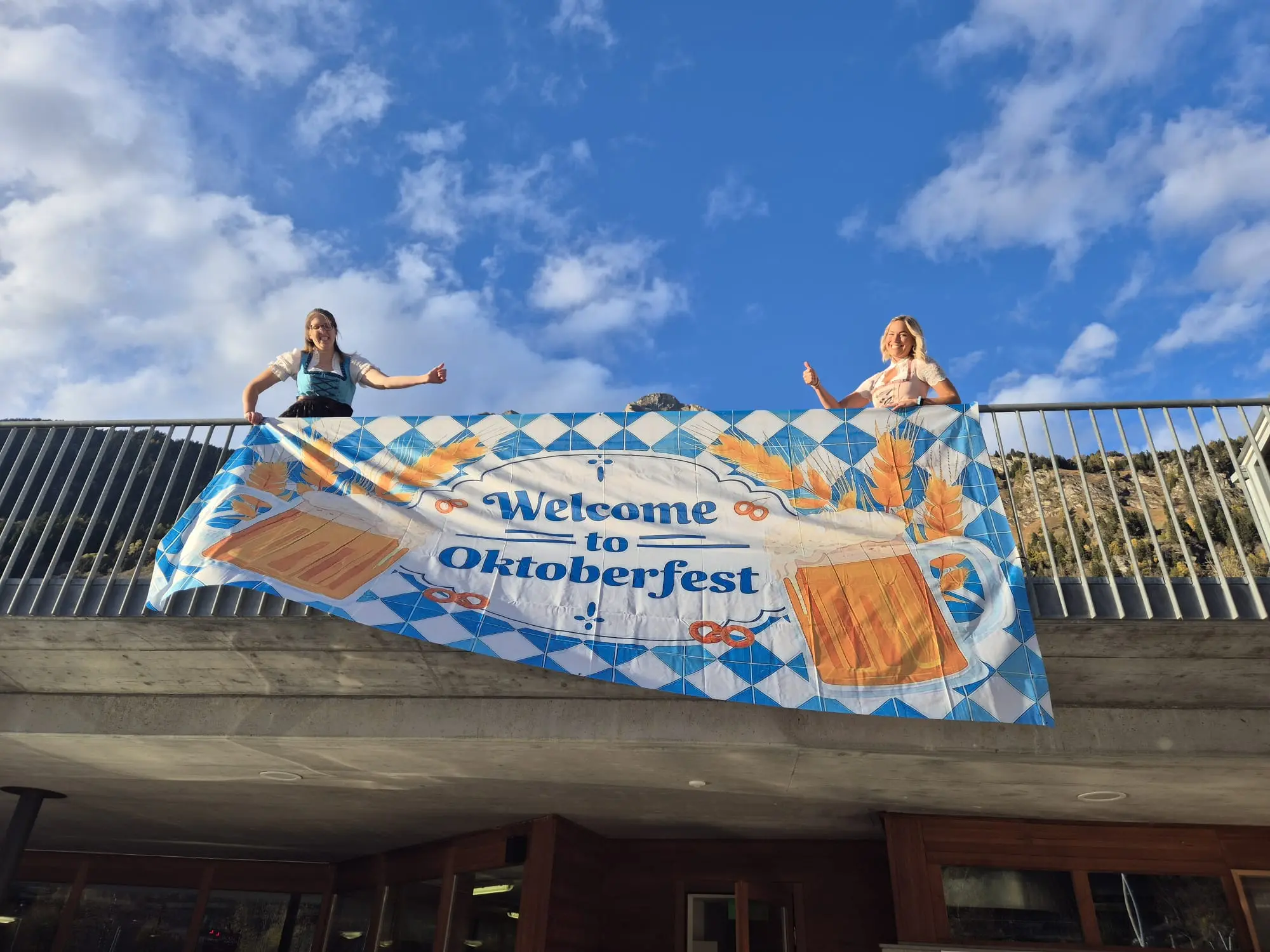 Due donne in abiti tradizionali festeggiano su un balcone, mostrando un grande striscione che dice "Welcome to Oktoberfest", decorato con boccali di birra e motivi blu e bianchi. Il cielo è sereno, con poche nuvole.