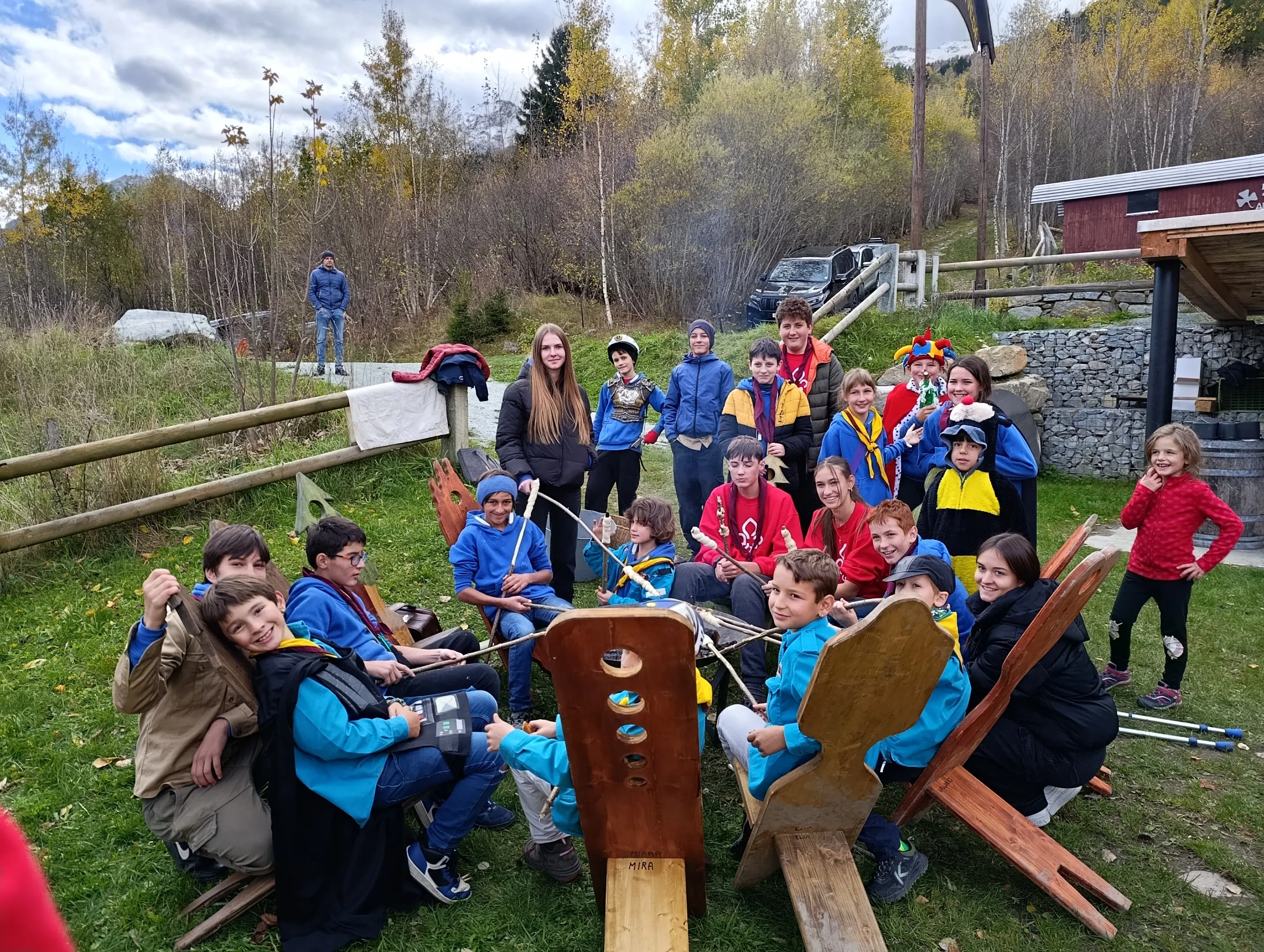 Gruppo di ragazzi e adulti riuniti all'aperto, sorridenti e seduti su sedie di legno, con attrezzature ludiche e colorate. Alcuni indossano costumi e accessori divertenti, circondati da un ambiente naturale autunnale.