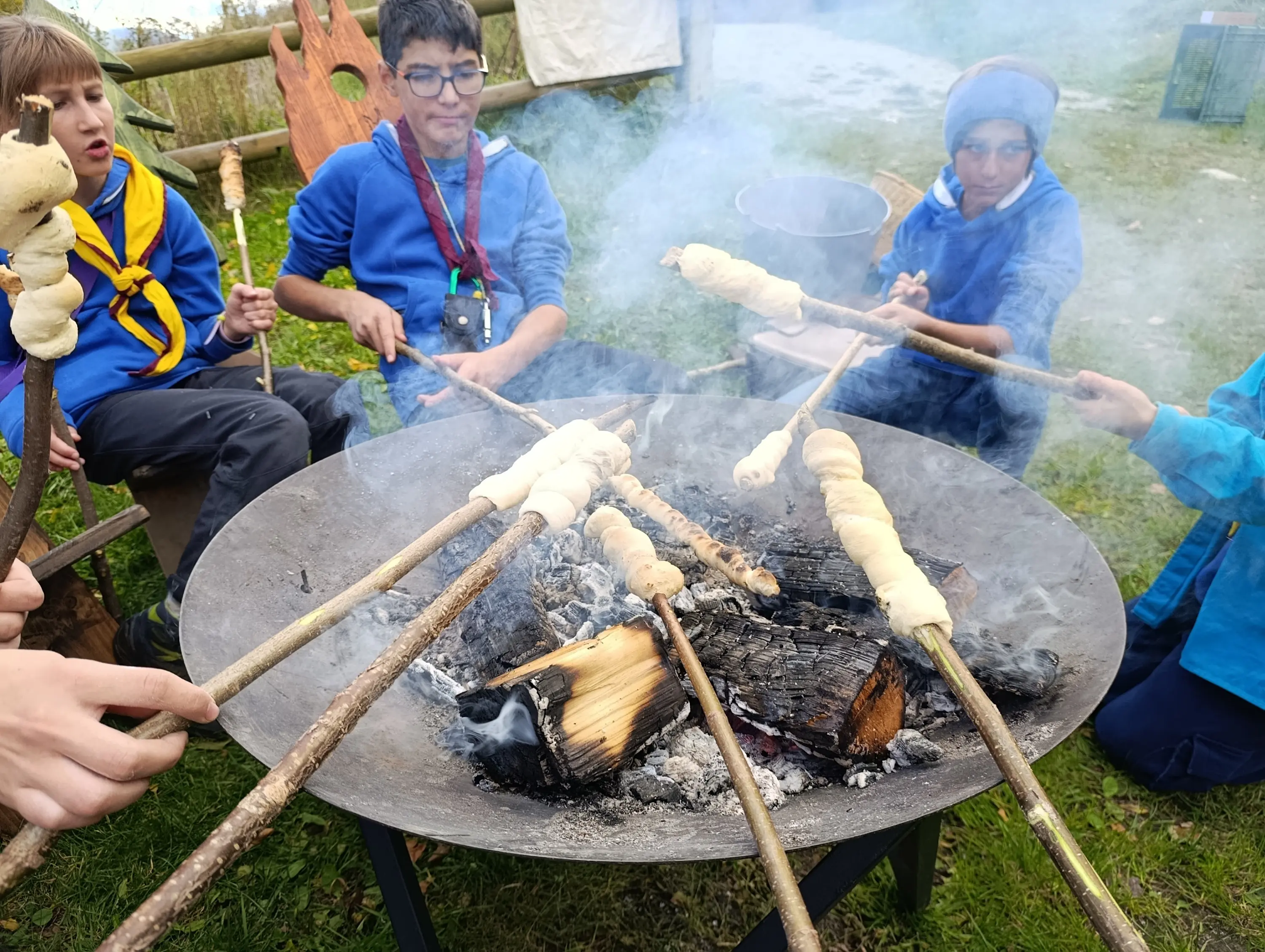 Un gruppo di ragazzi seduti intorno a un falò mentre cuociono impasti attaccati a bastoni. C'è fumo che si alza dal fuoco e un'atmosfera di convivialità.