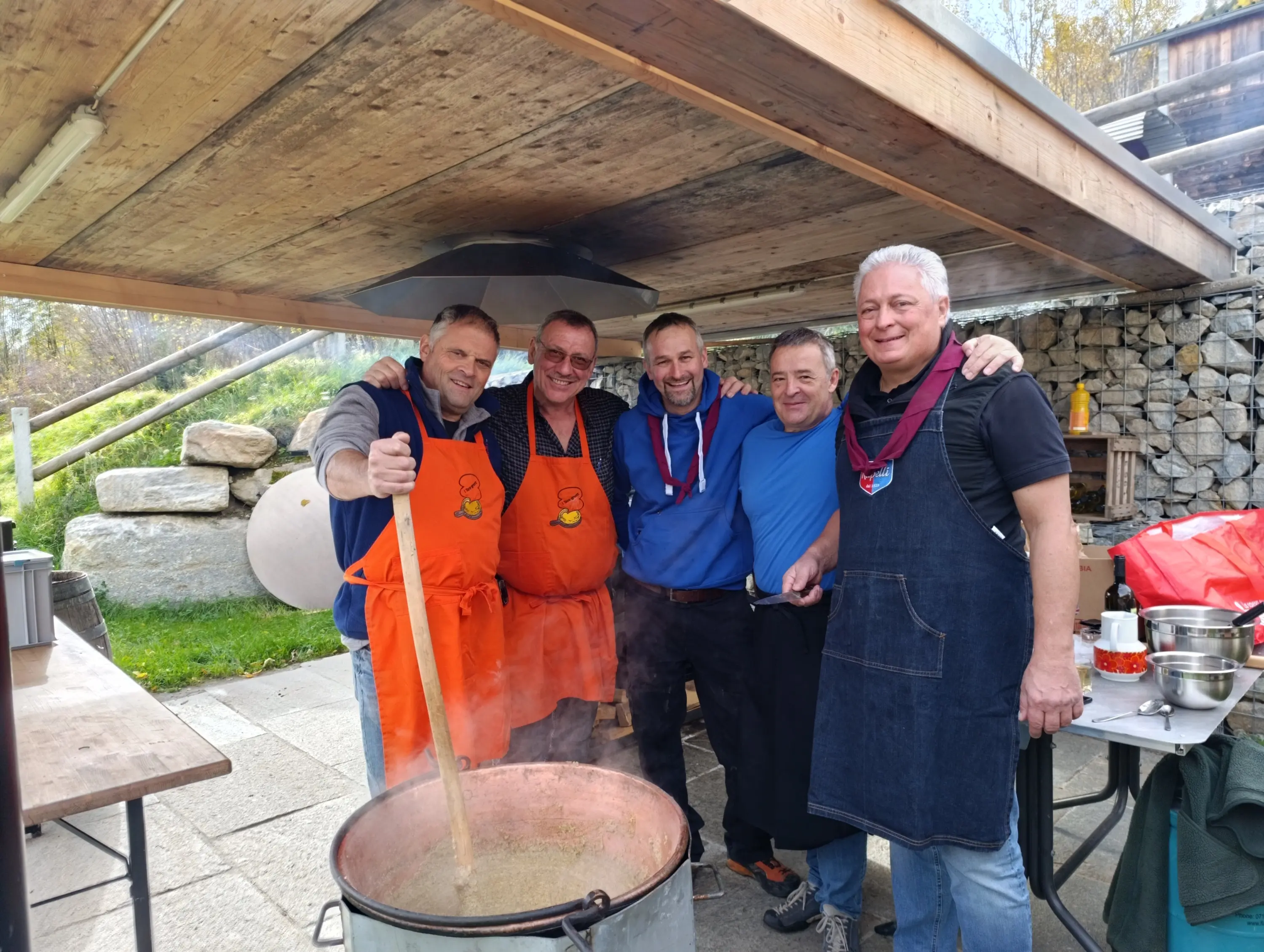 Cinque uomini sorridenti indossano grembiuli da cucina, mentre mescolano un grande pentolone su una tavola all'aperto. Sullo sfondo, una struttura di legno e sassi con un'atmosfera rustica.