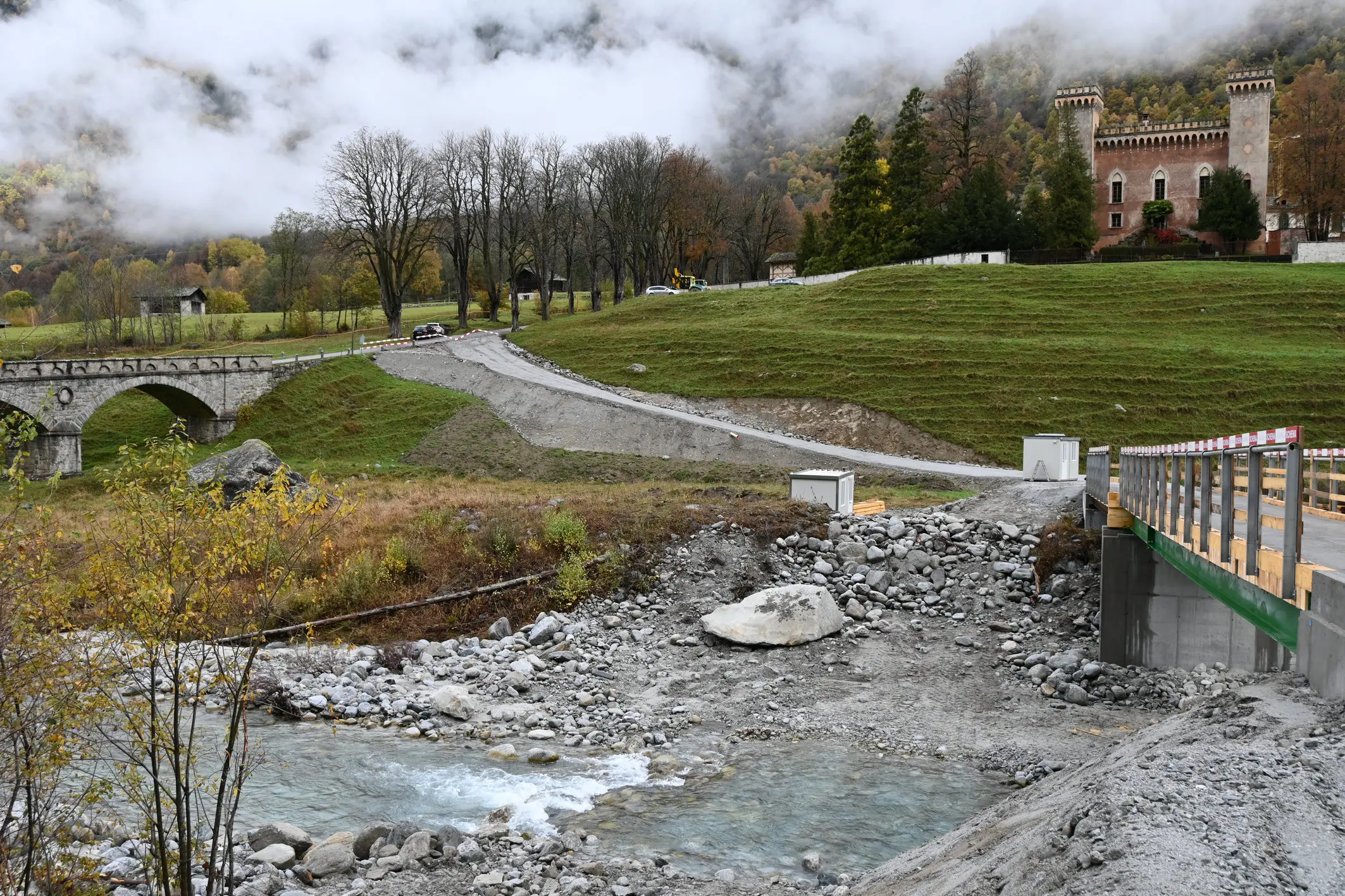 Una vista di un paesaggio montano con un castello sullo sfondo. Un ponte in pietra attraversa un fiume che scorre tra le rocce. Gli alberi con foglie autunnali si trovano nei dintorni, mentre le nuvole coprono par