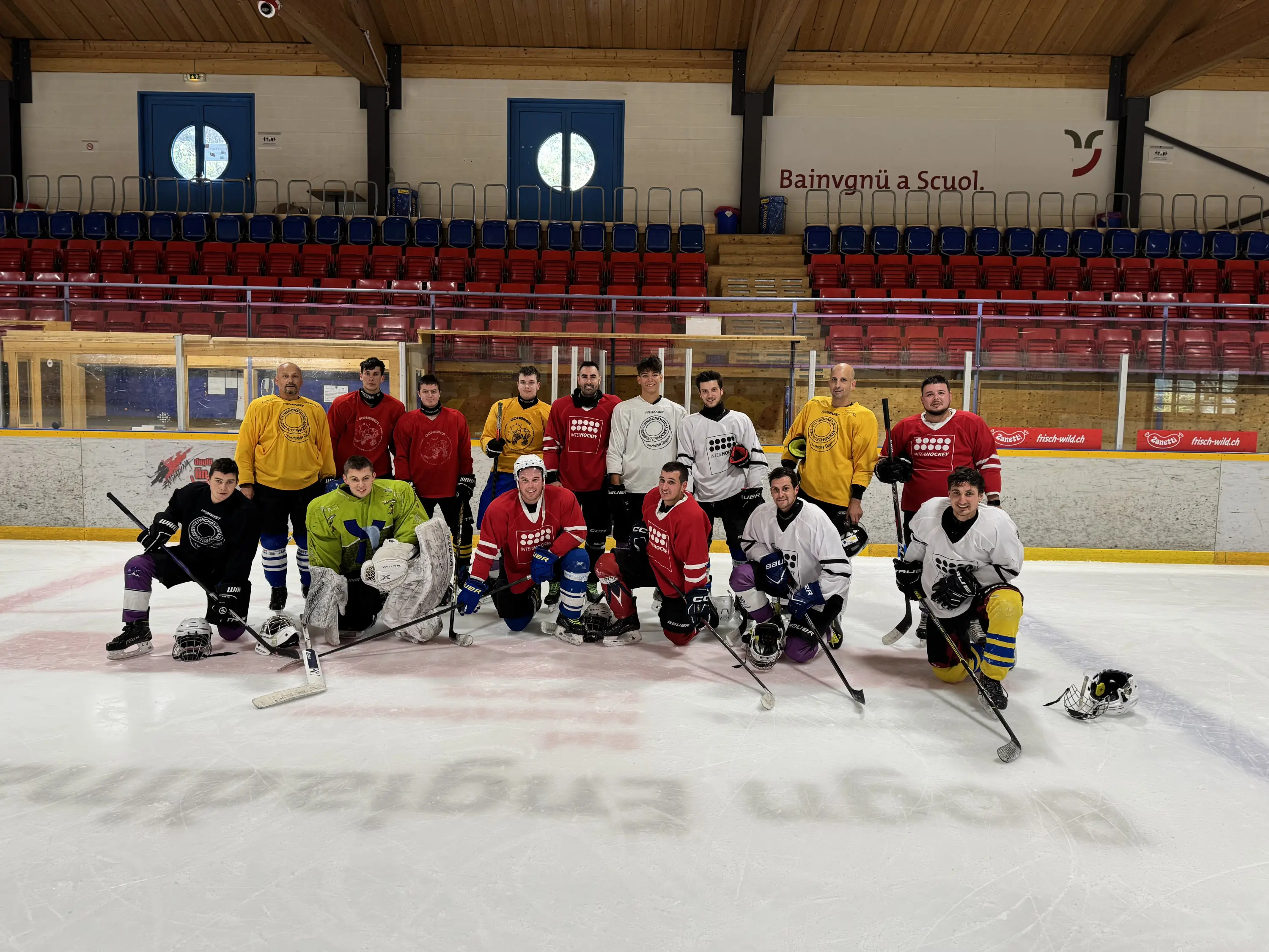 Un gruppo di giocatori di hockey su ghiaccio posa sul ghiaccio di un'arena, indossando maglie colorate: giallo, rosso, bianco e nero. Gli atleti, divisi in squadre, sorridono e mostrano le attrezzature da