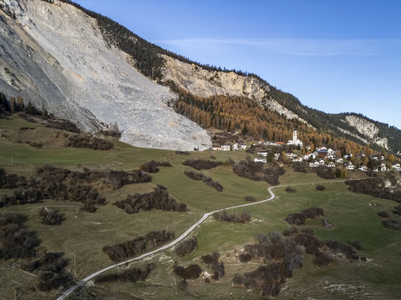 Una vista panoramica di una valle con un'insenatura di frana, circondata da alberi autunnali. In lontananza si vede un piccolo villaggio con un campanile e un sentiero che si snoda tra i prati verdi.