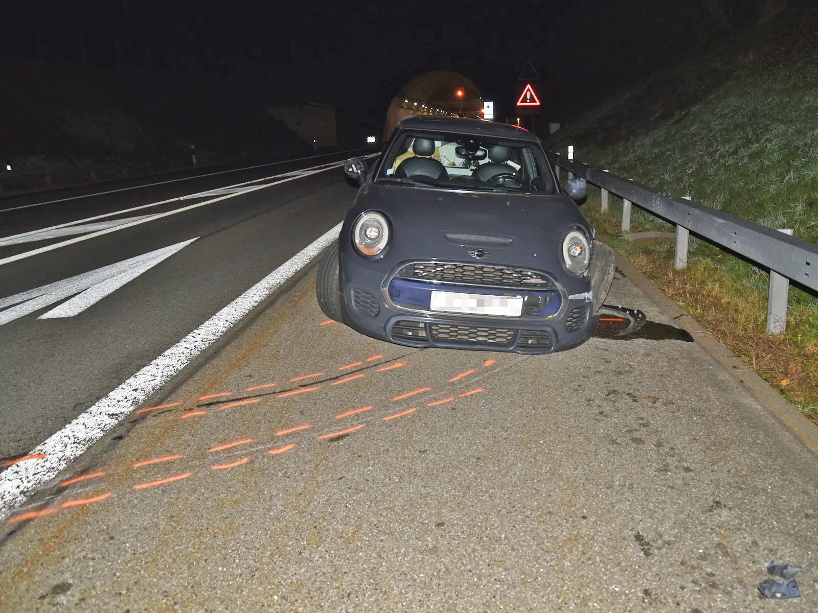 Un’auto nera parcheggiata sul lato di una strada, con un tunnel in lontananza. L'auto sembra essere danneggiata, con segni di frenata visibili sull'asfalto. Un cartello di avvertimento è presente nella zona.