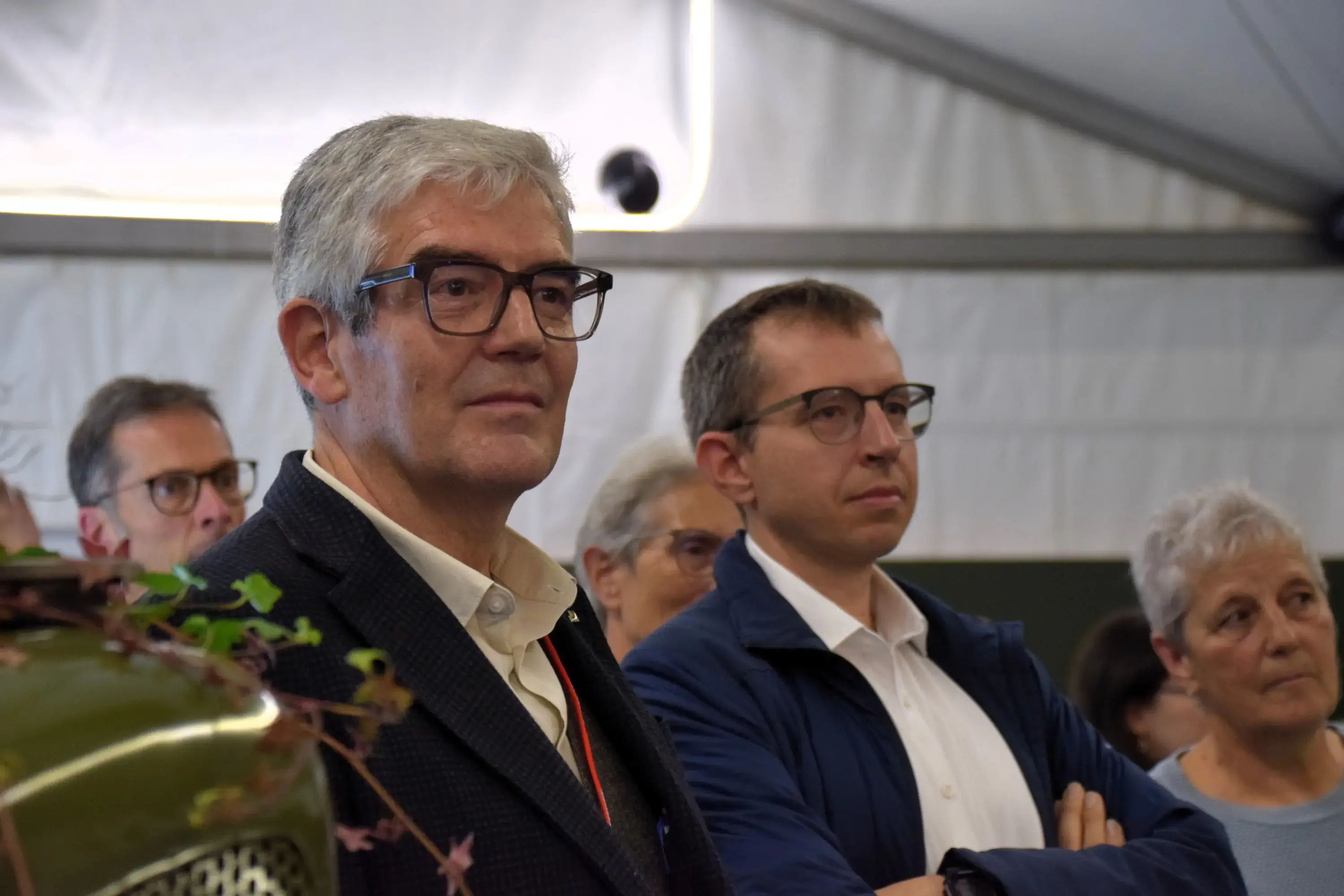 Gruppo di persone in un evento. Due uomini con occhiali osservano attentamente, mentre una donna anziana a destra ascolta. Lo sfondo è un ambiente interno con tende.