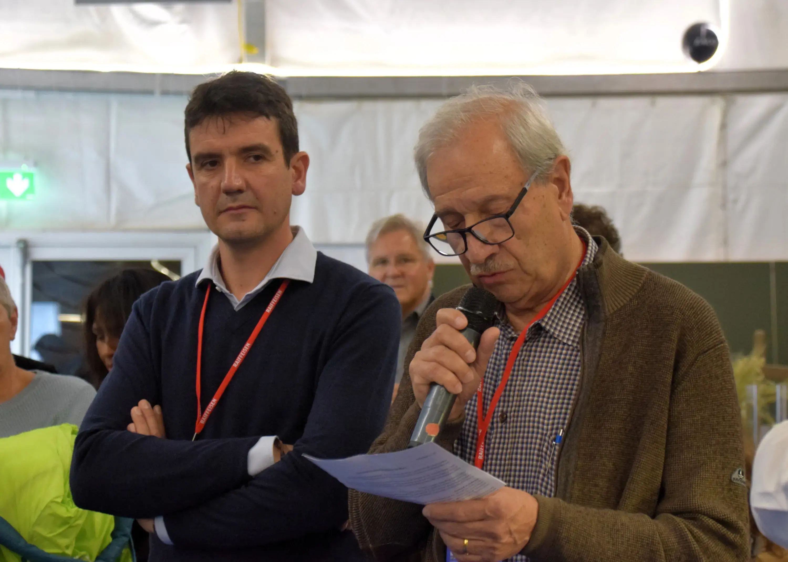 Un uomo anziano legge un discorso al microfono, mentre un uomo più giovane lo ascolta con attenzione sullo sfondo. Entrambi indossano maglioni e collane identificative. L'ambiente sembra essere una sala riunioni.