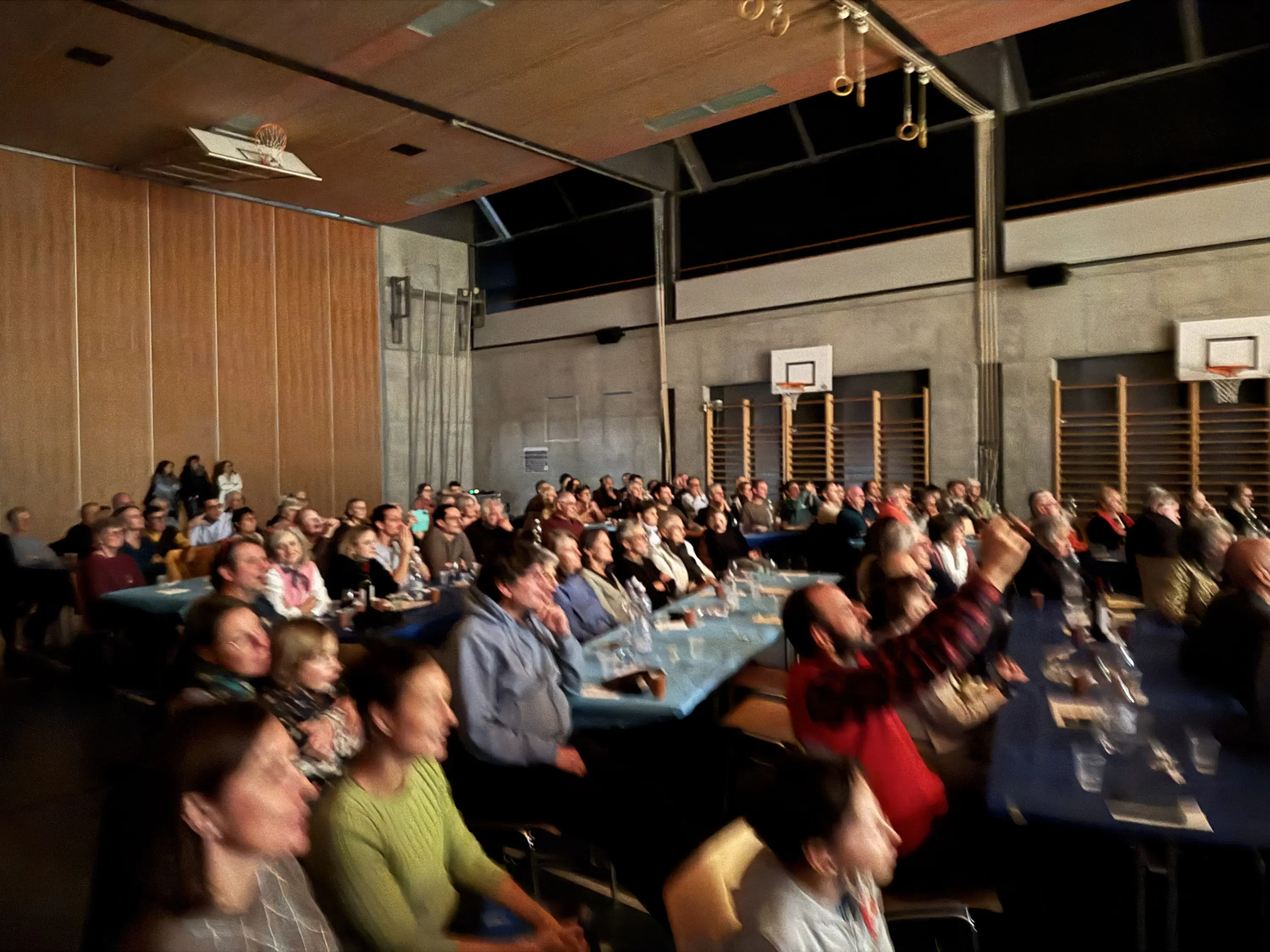 Sala affollata con pubblico che assiste a un evento. Ci sono persone di diverse età, alcune sorridono e altre applaudono. Tavoli blu con bicchieri e bottiglie. Sullo sfondo, le attrezzature sportive e un palco.