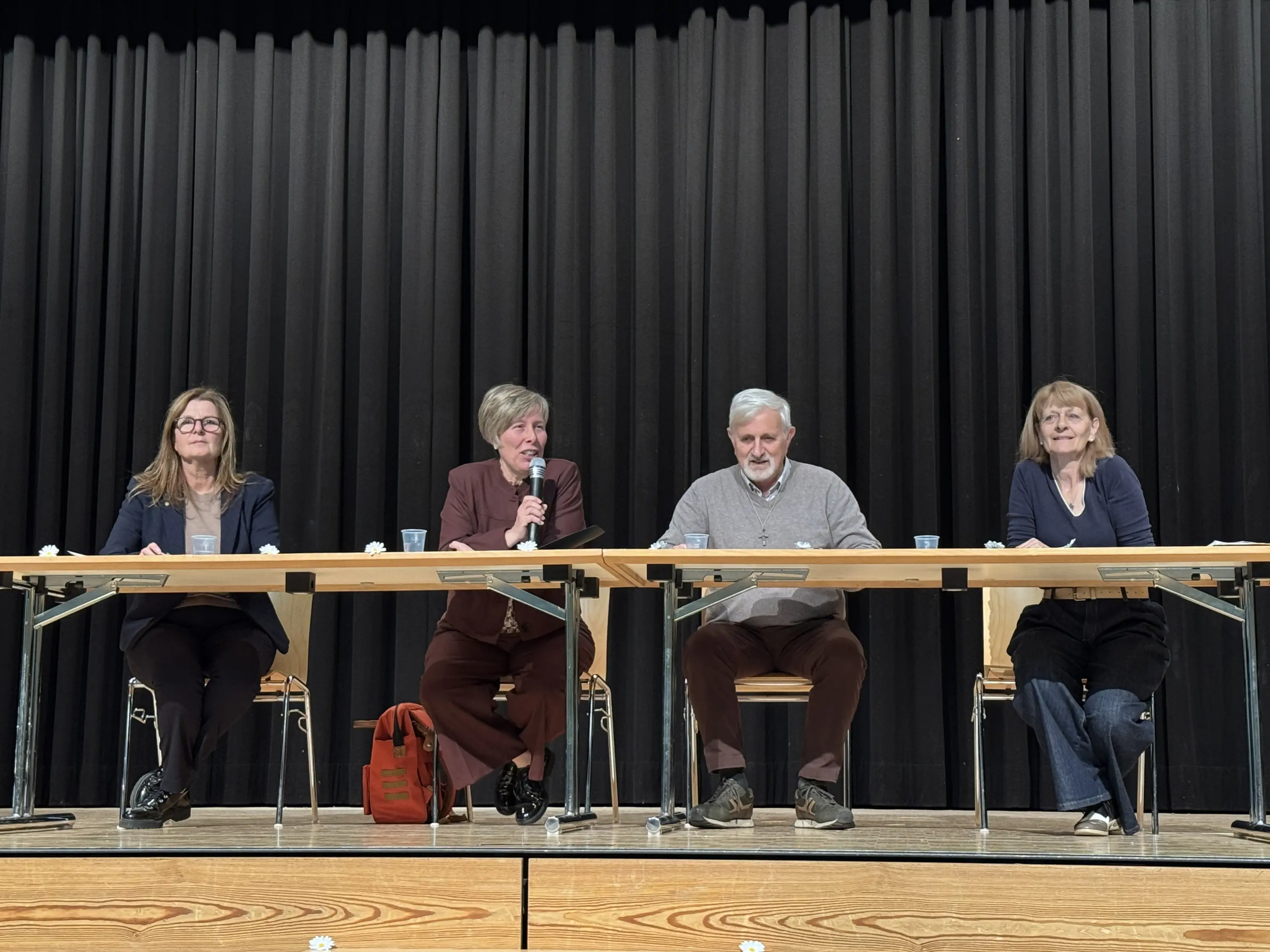 Quattro relatori siedono a un tavolo su un palco. Tutti sorridono e stanno partecipando a un evento, con microfoni e bicchieri d'acqua davanti a loro. Il fondale è una parete nera drappeggiata.