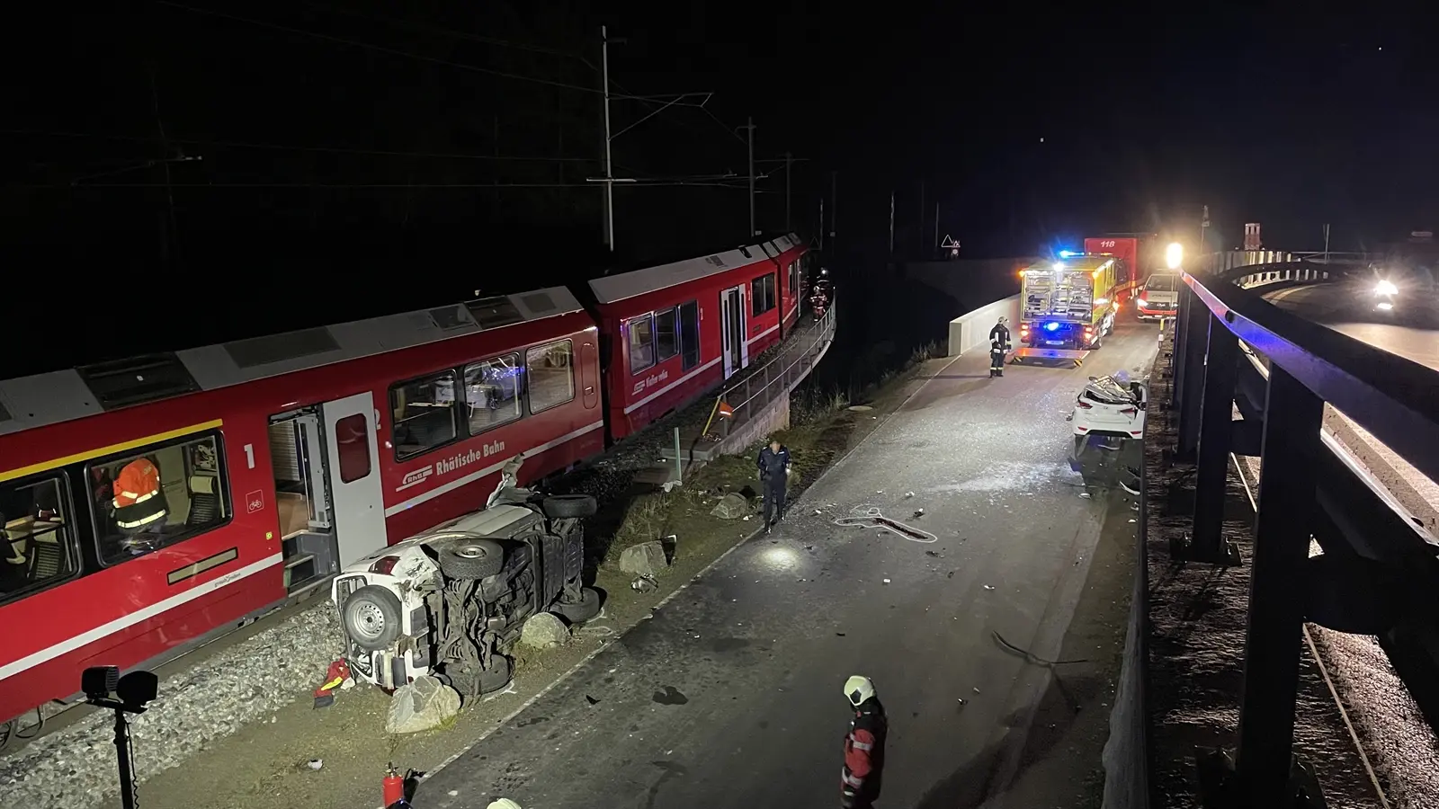 Incidente ferroviario notturno con un treno deragliato su un binario. I soccorritori lavorano sulle scene, mentre un'auto ribaltata si trova lungo la strada adiacente. Luci di emergenza lampeggianti illuminano l'area.