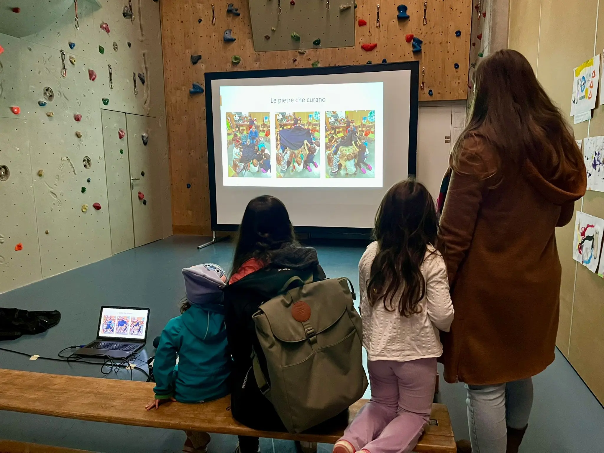 Due adulti e due bambini osservano una presentazione su uno schermo in una sala con pareti da arrampicata. Un computer portatile è aperto su una panchina di legno.