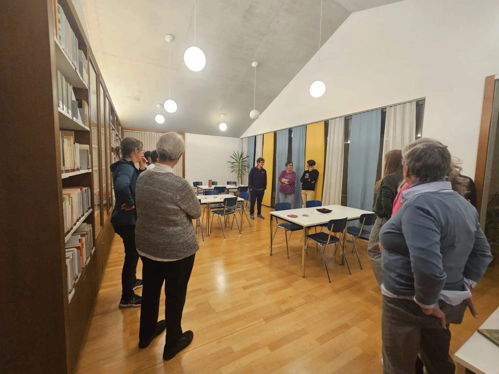 Un gruppo di persone si trova in una stanza luminosa e accogliente, con tavoli e sedie. Alcuni partecipanti stanno conversando mentre altri ascoltano, circondati da scaffali di libri. La stanza ha finestre ampie e decorazioni nei toni del g