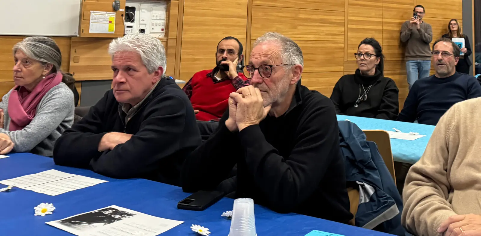 Un gruppo di persone sedute a un tavolo con una tovaglia azzurra, ascoltando attentamente. Un uomo con occhiali è al centro, mentre gli altri sembrano interessati. Sul tavolo ci sono fogli e bicchieri.