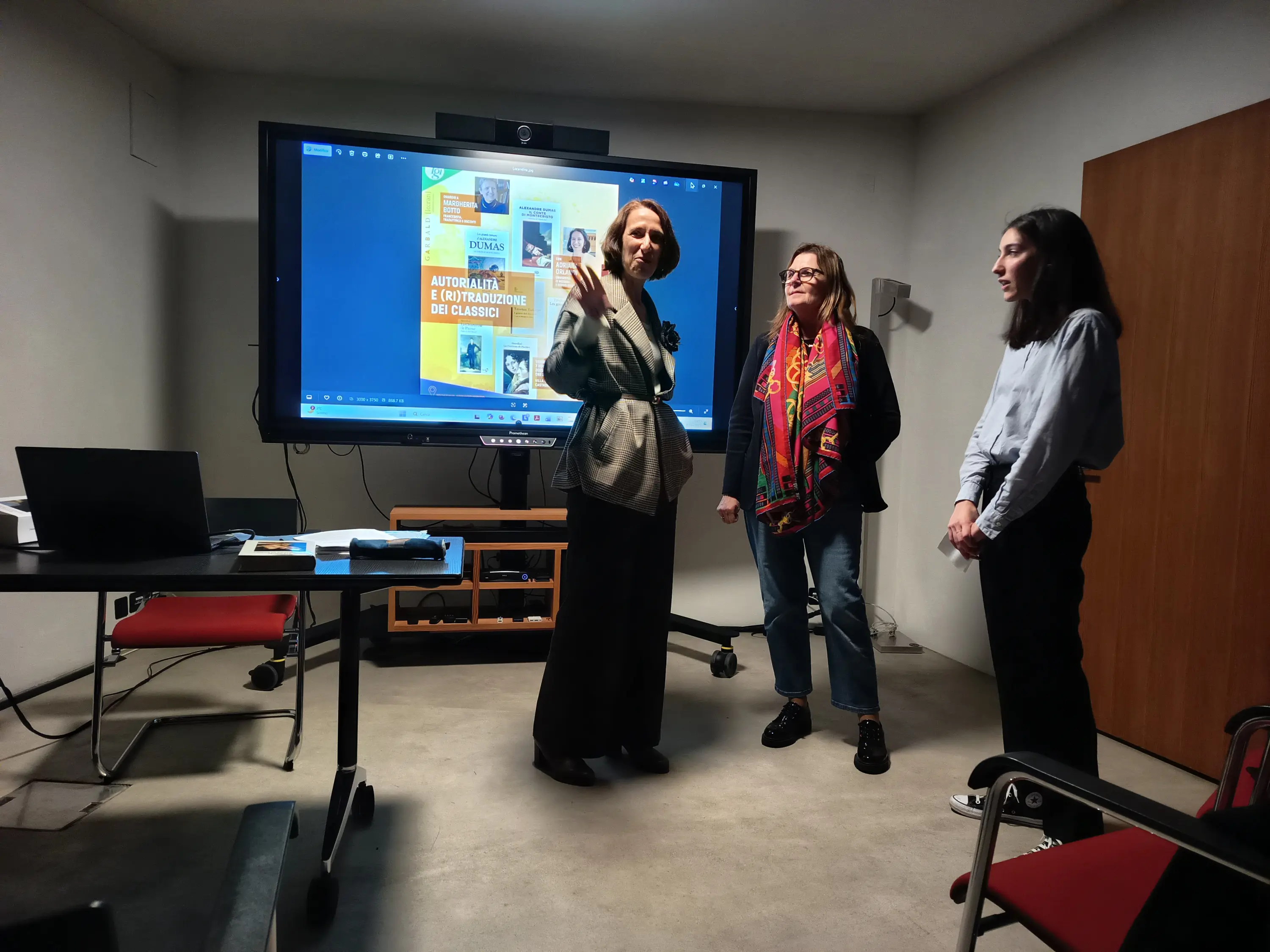 Tre donne in un ambiente di lavoro, con un grande schermo dietro di loro che mostra un poster. Due donne sono in piedi mentre una sta illustrando qualcosa. Una ha una giacca grigia e l'altra indossa una sciarpa colorata. Una terza donna osserva,