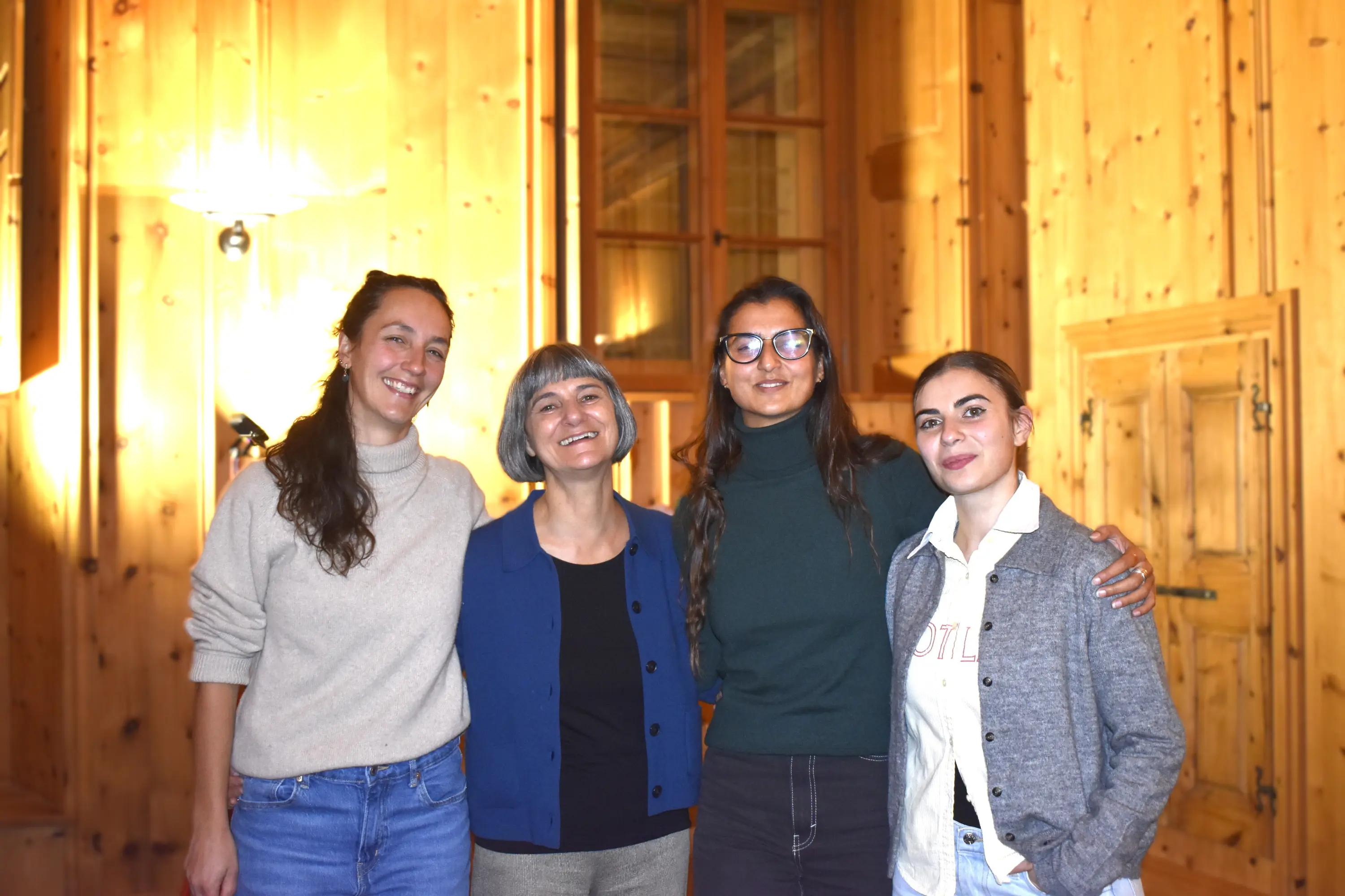 Quattro donne sorridenti posano insieme in una stanza con pareti di legno. La luce calda illumina l'ambiente, creando un'atmosfera accogliente.