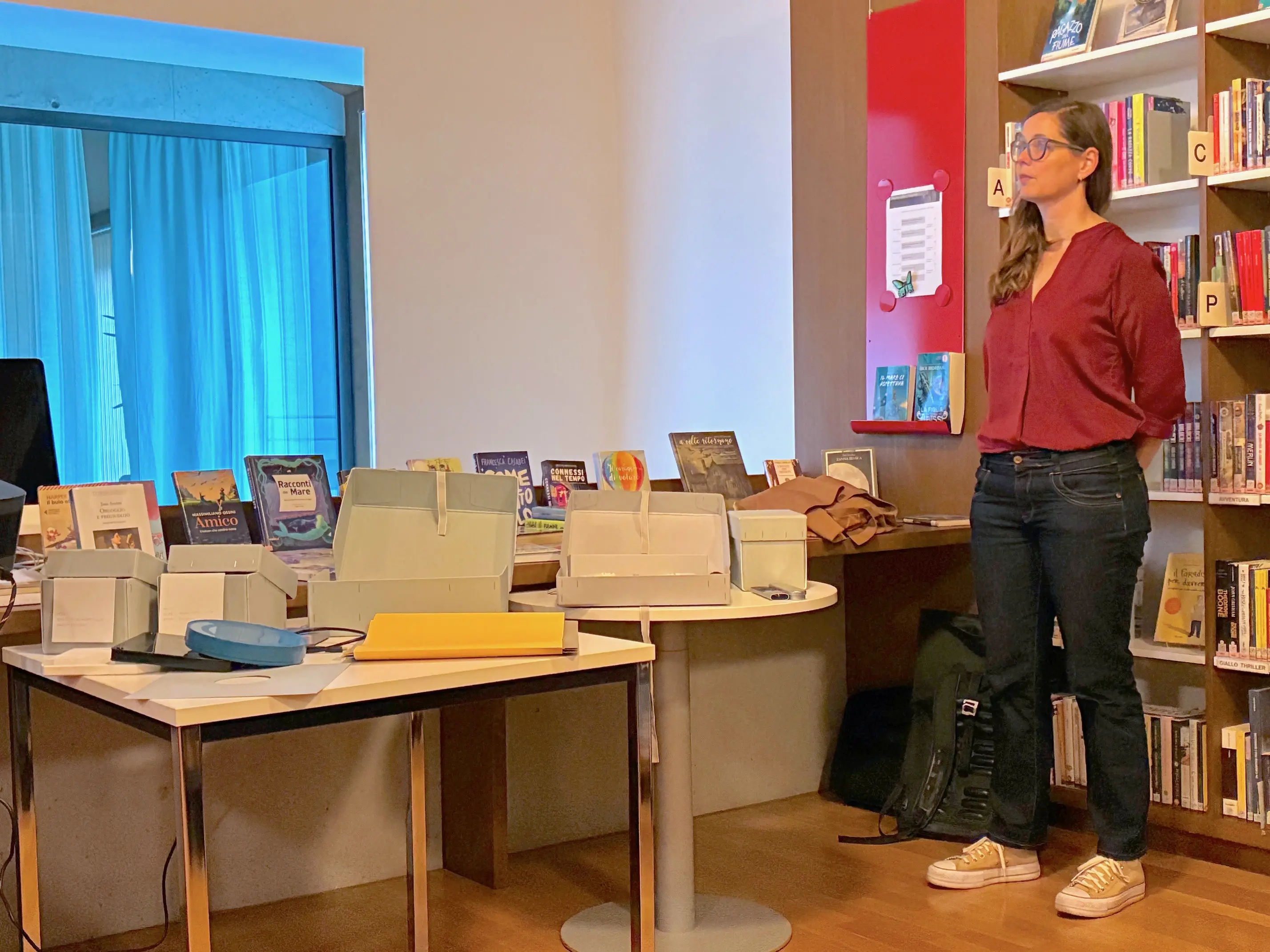 Una donna in piedi accanto a un tavolo con scatole e libri in una biblioteca. Sullo sfondo si vedono scaffali di libri e una finestra con luce blu.