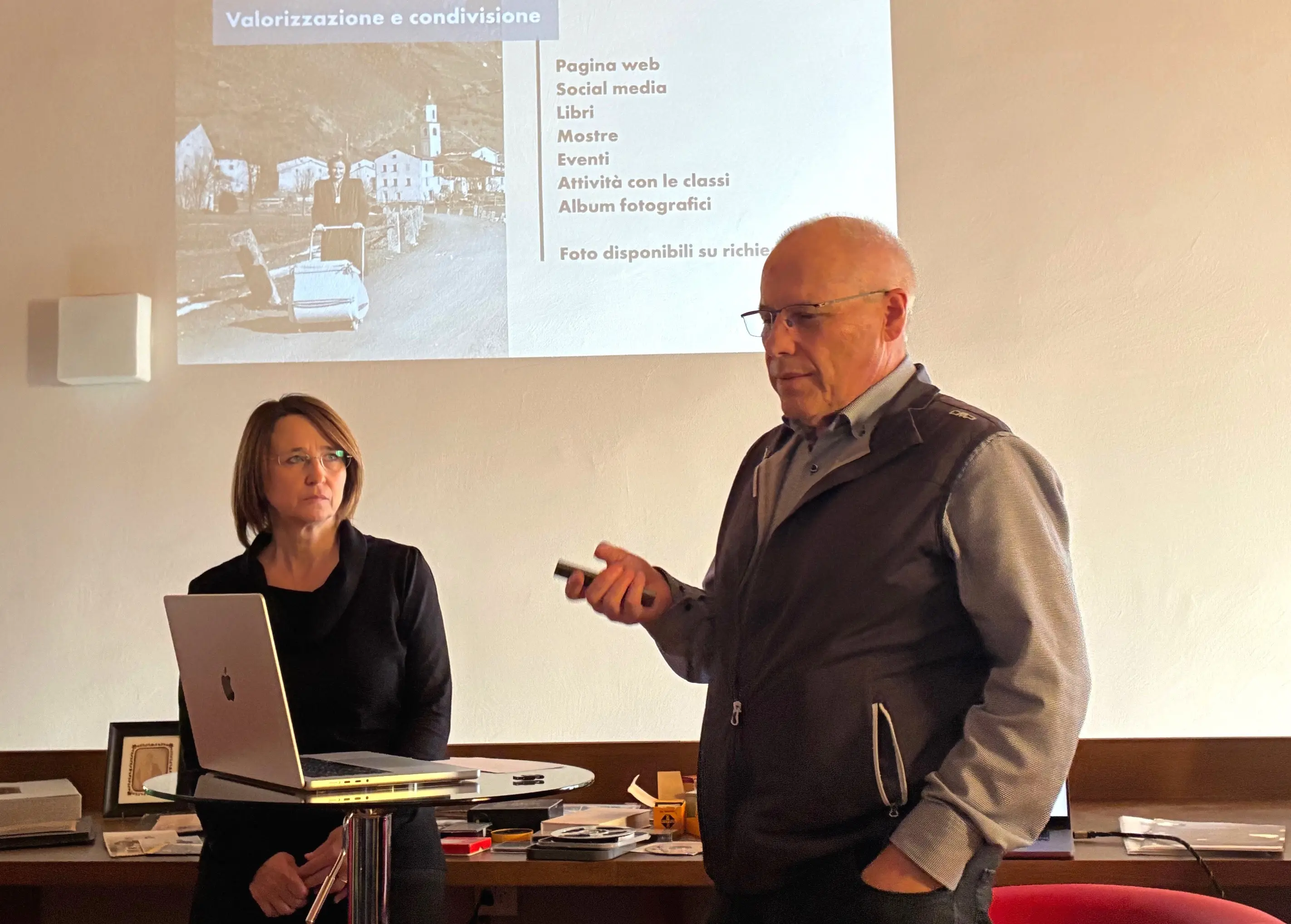 Due persone stanno presentando un progetto. Un uomo anziano, con occhiali e un gilet, parla tenendo in mano un dispositivo. Una donna, di fronte a lui, ascolta attentamente. Sullo sfondo, si vede una proiezione con testi riguardanti web