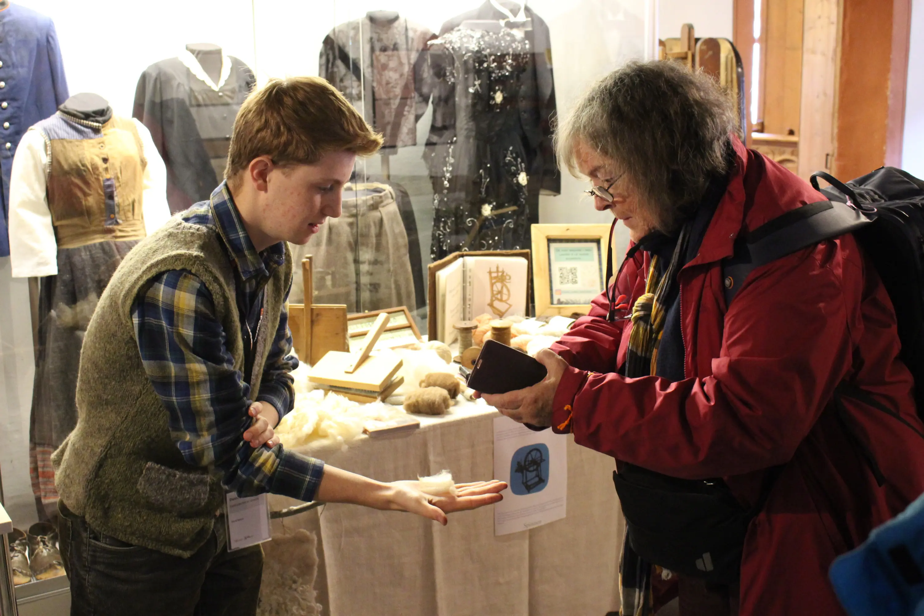 Un giovane in maglione mostra della lana a un visitatore più anziano, mentre vari oggetti storici e abbigliamento sono esposti in una vetrina dietro di loro.