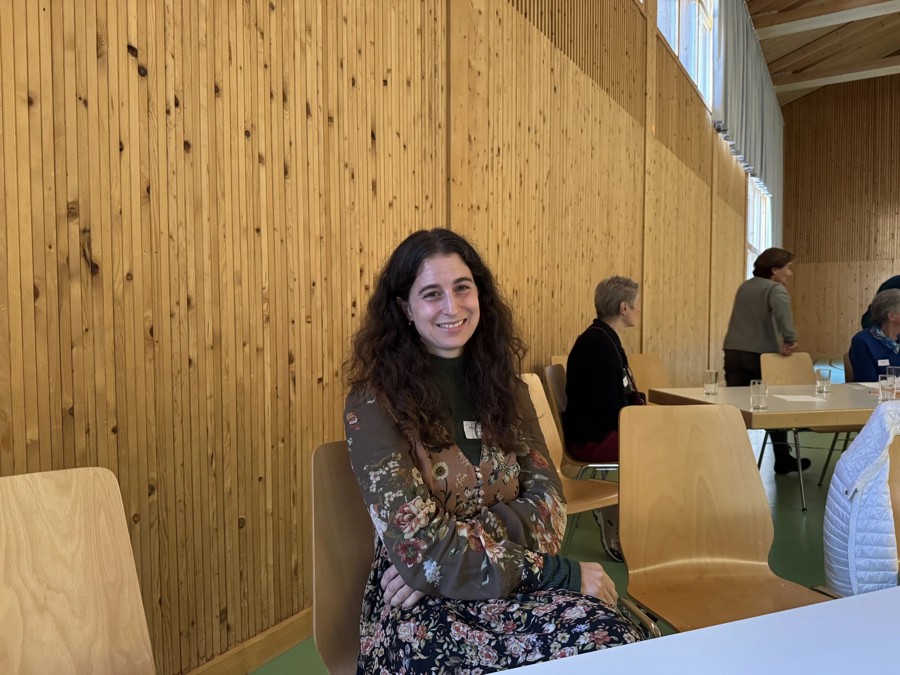 Una giovane donna con capelli ricci e lunghi sorride mentre è seduta in una sala con pareti di legno. Indossa una giacca floreale e si trova tra altre persone.