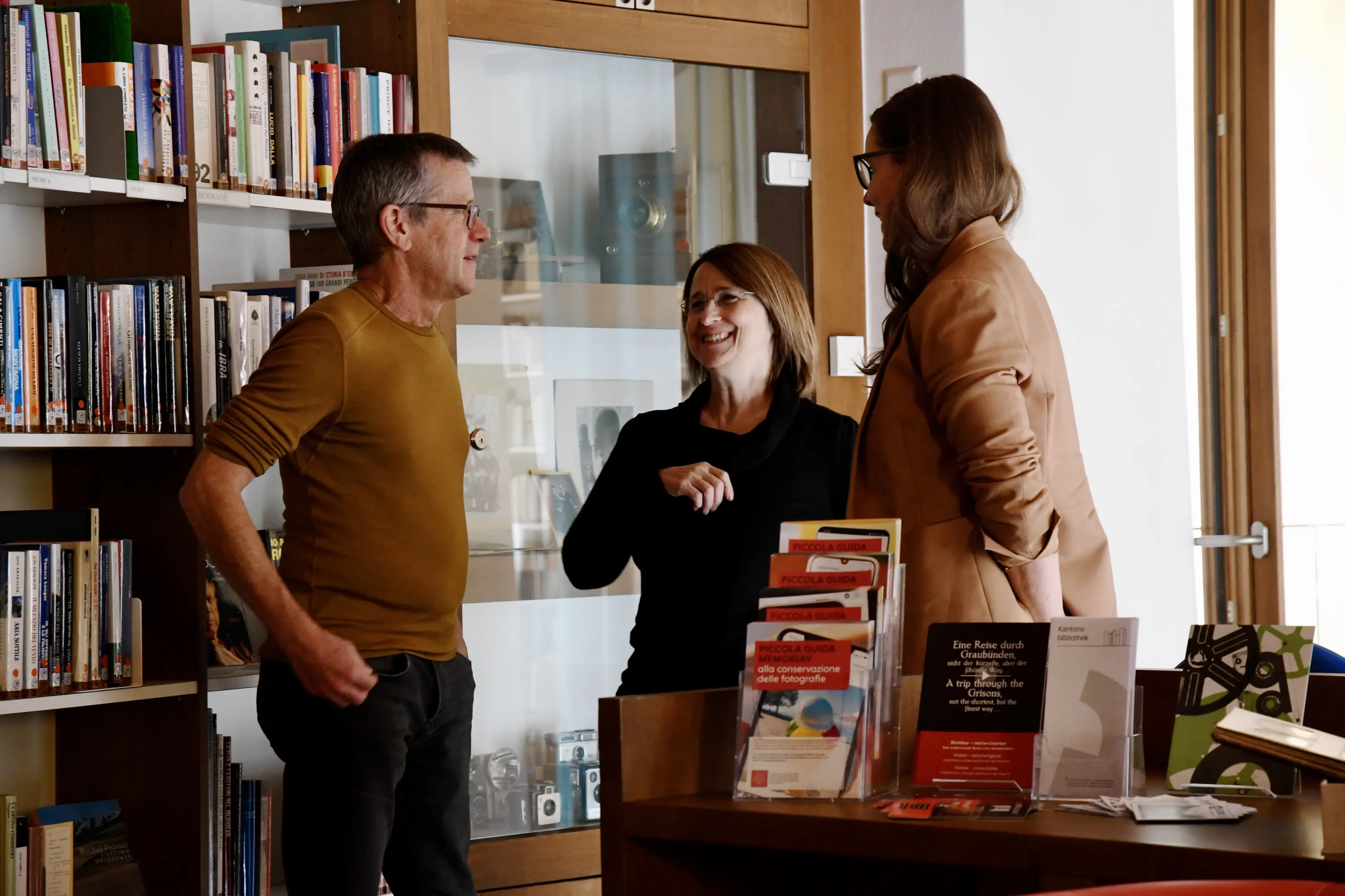 Due persone sorridenti sono in conversazione con un uomo in una biblioteca. Sullo sfondo si vedono scaffali pieni di libri e sulla scrivania ci sono vari opuscoli e materiali informativi.